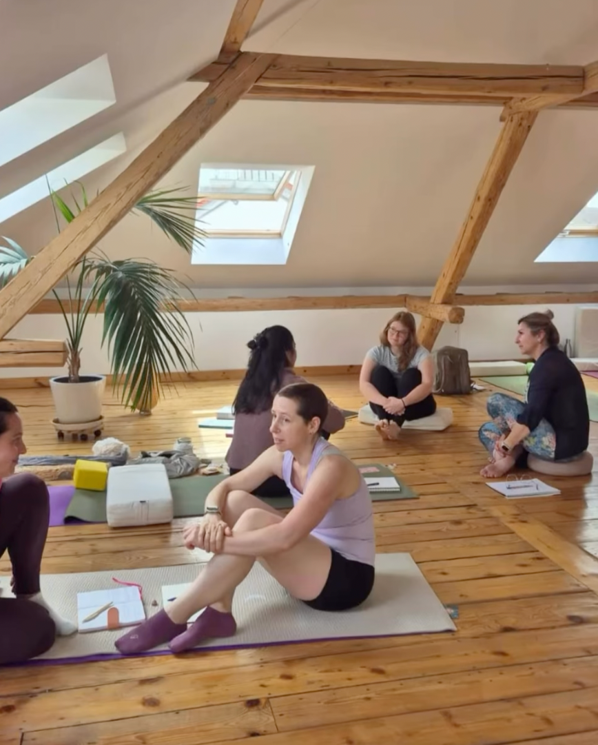 Menschen sitzen auf Yogamatten in einem Raum mit Holzboden und Dachfenstern, unterhalten sich, während sie Yoga- oder Meditationsübungen machen.