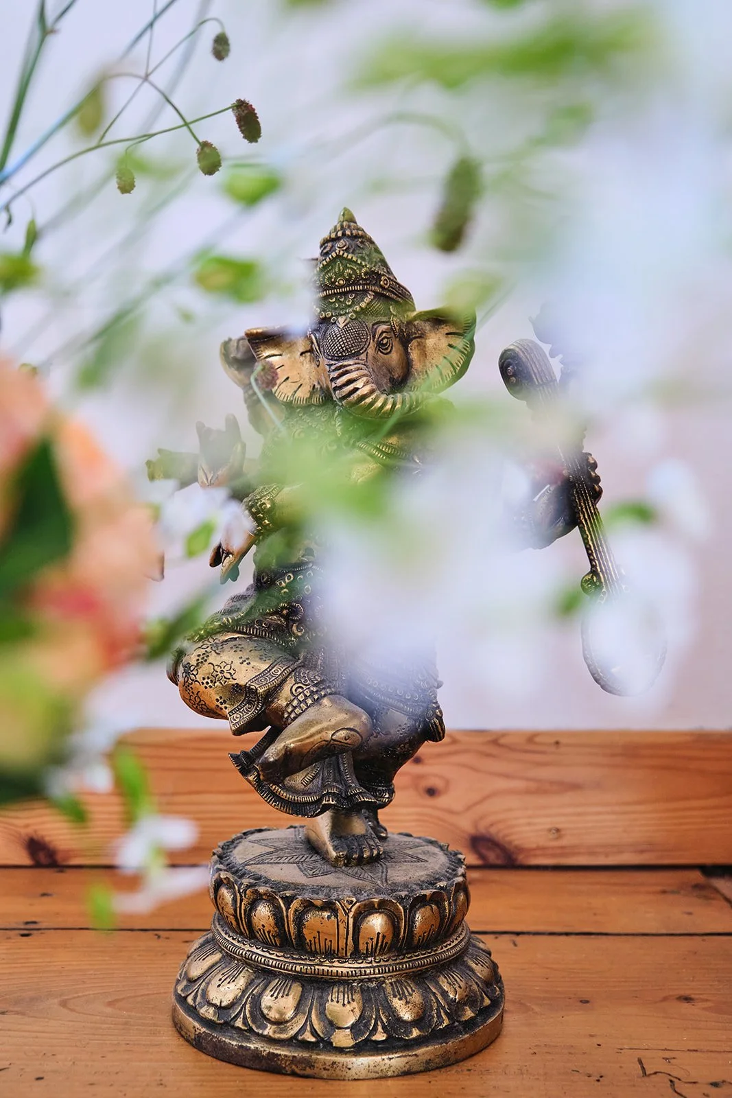 Bronzefarbene Statue eines tanzenden Hindu-Gottes, vermutlich Ganesha, auf einem Holzboden, umgeben von grünen Blättern und weißen Blumen.