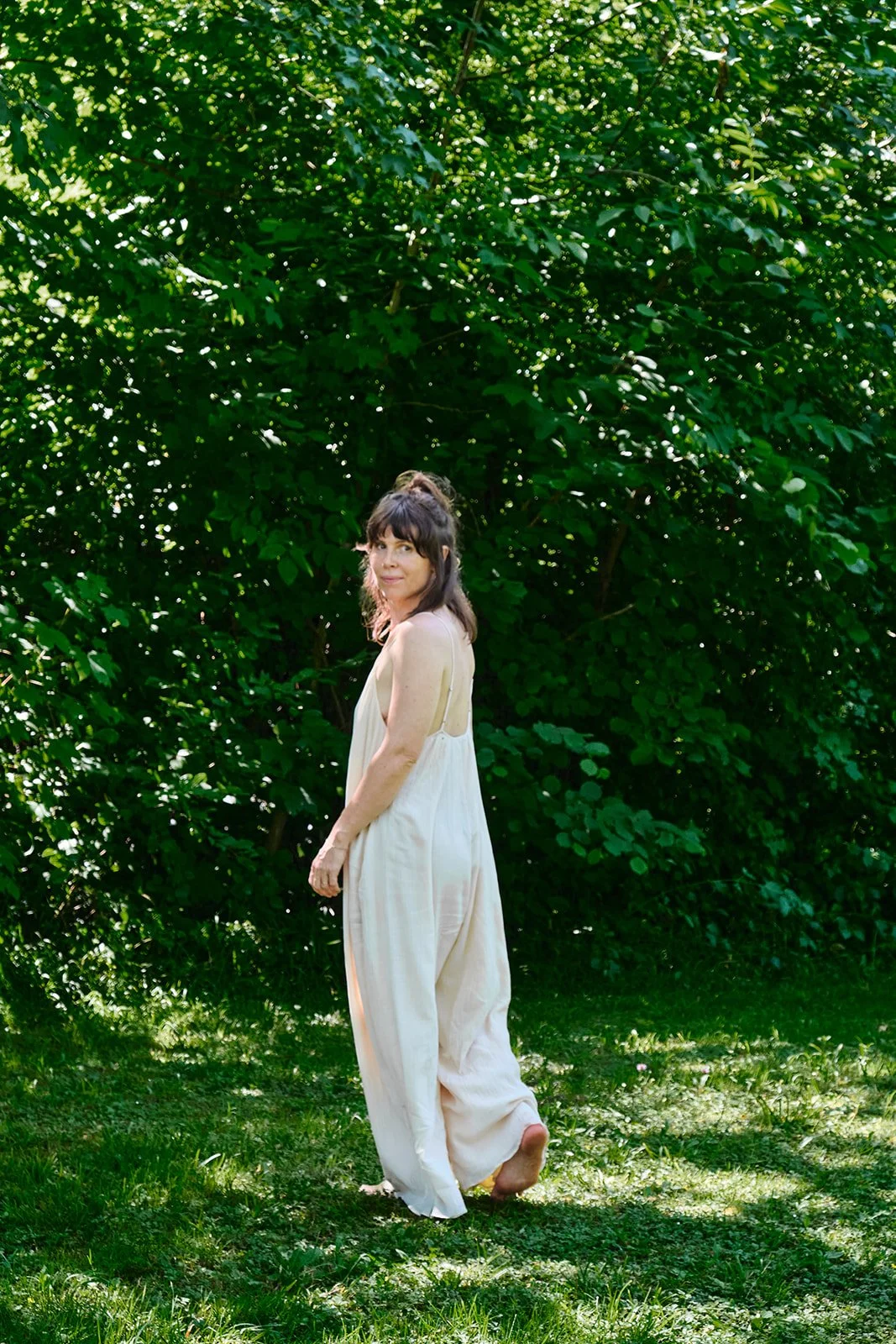 Eine Frau in einem weißen Kleid steht barfuß auf dem Rasen vor einem dichten, grünen Busch im Freien.