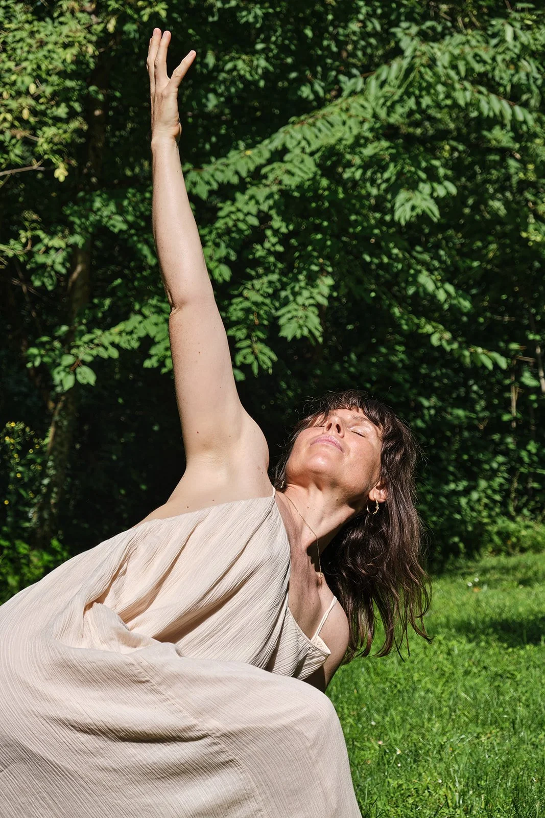 Frau im Freien, die sich in einer Yoga- oder Meditationspose befindet, mit Blick nach oben und einem Baum im Hintergrund.