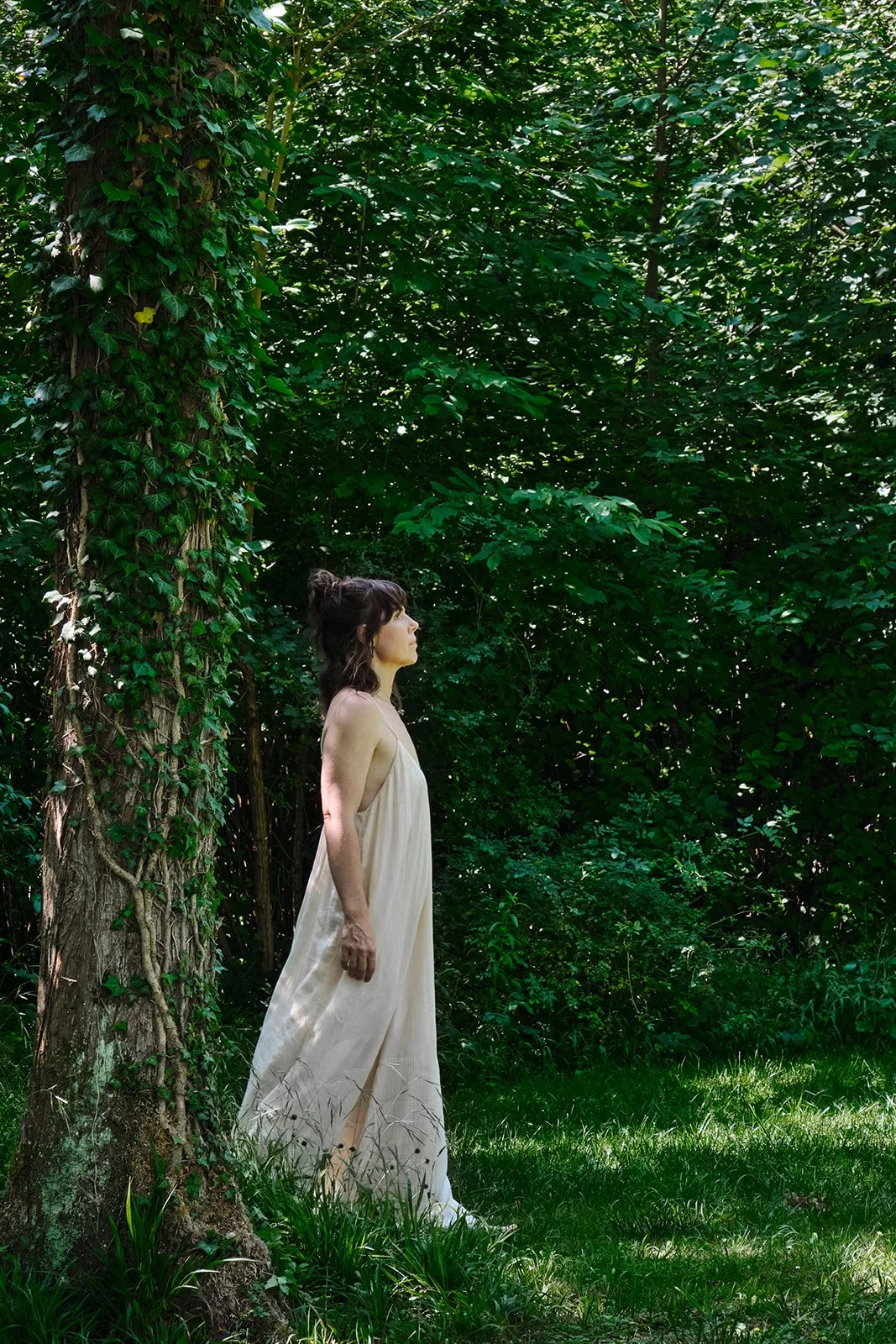 Eine Frau in einem weißen Kleid steht an einem Baum in einem dichten grünen Wald, schaut nach links und wirkt nachdenklich.