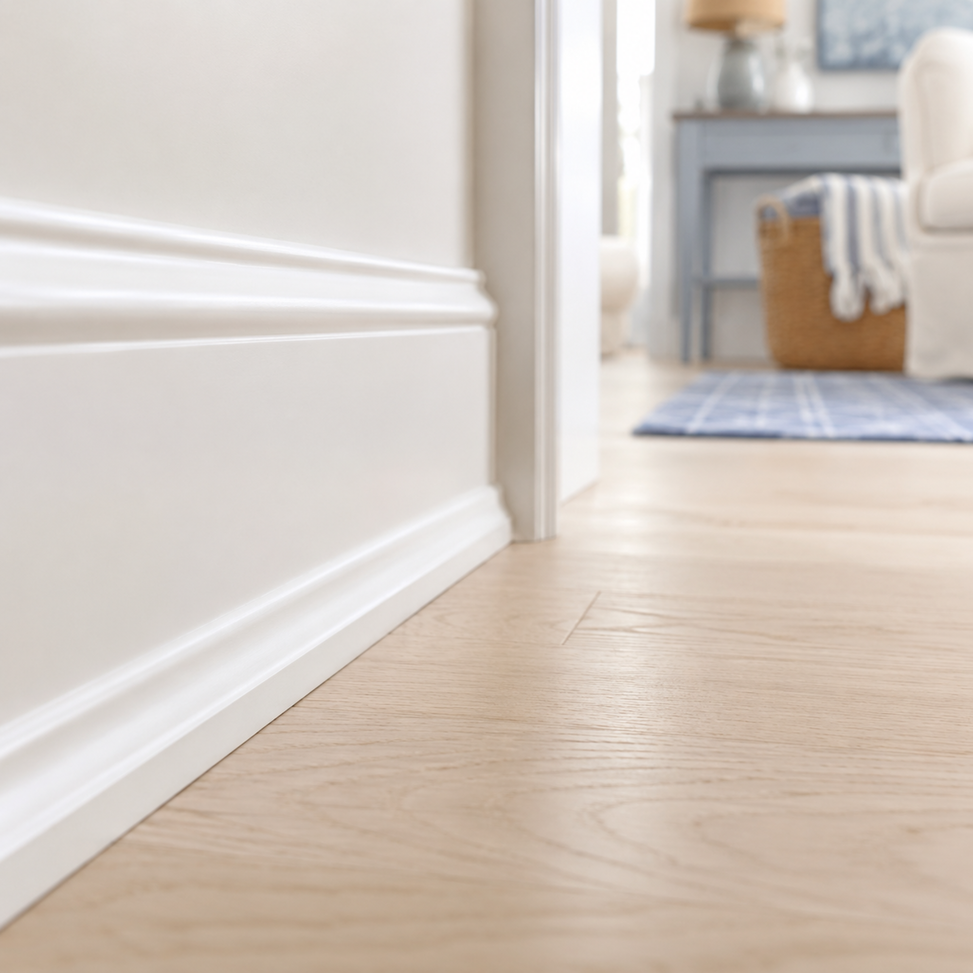 Close-up of a white baseboard along a wooden floor in a room, with part of a hallway in the background showing furniture and a rug.