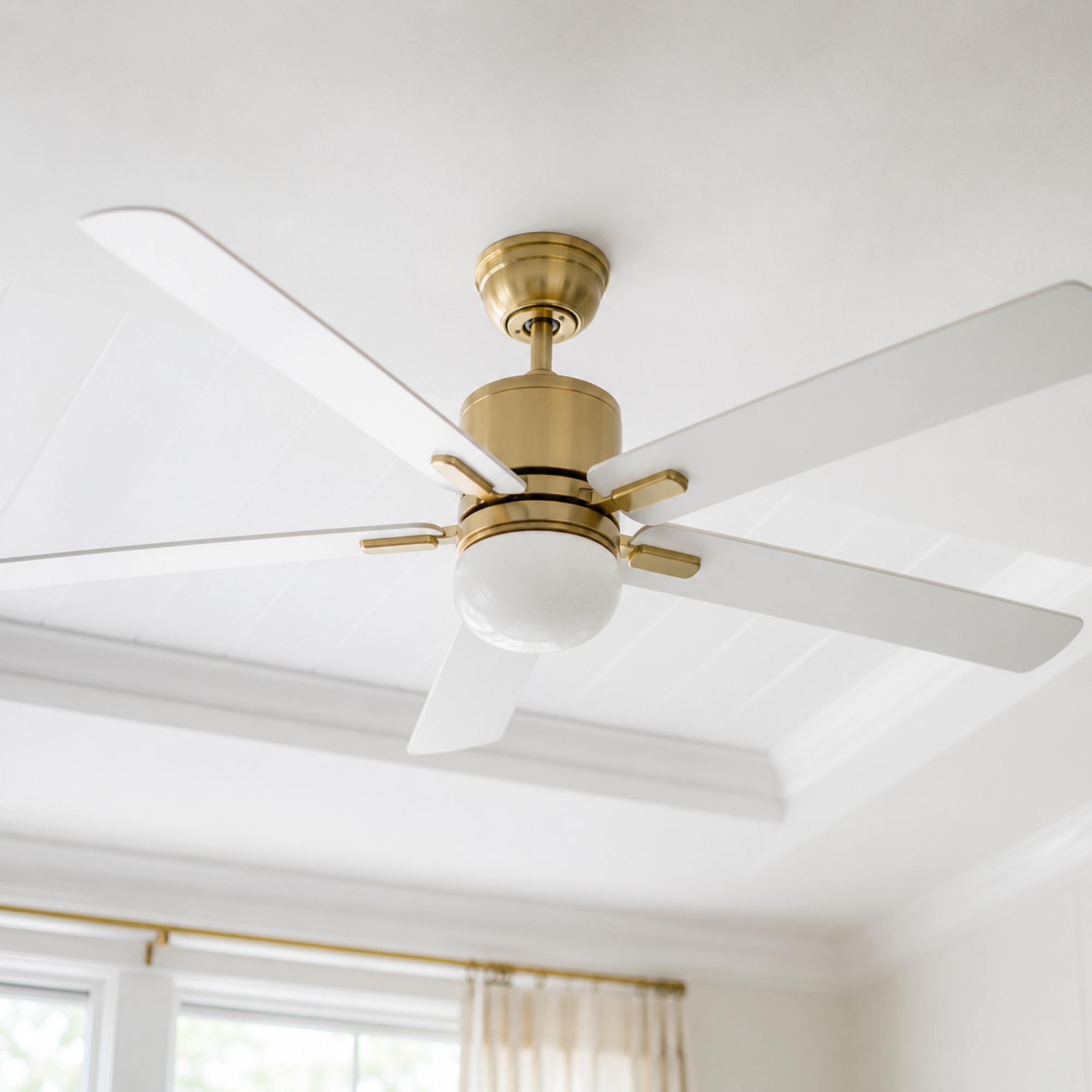 Ceiling fan with gold accents in a room with a white ceiling and large window with curtains.