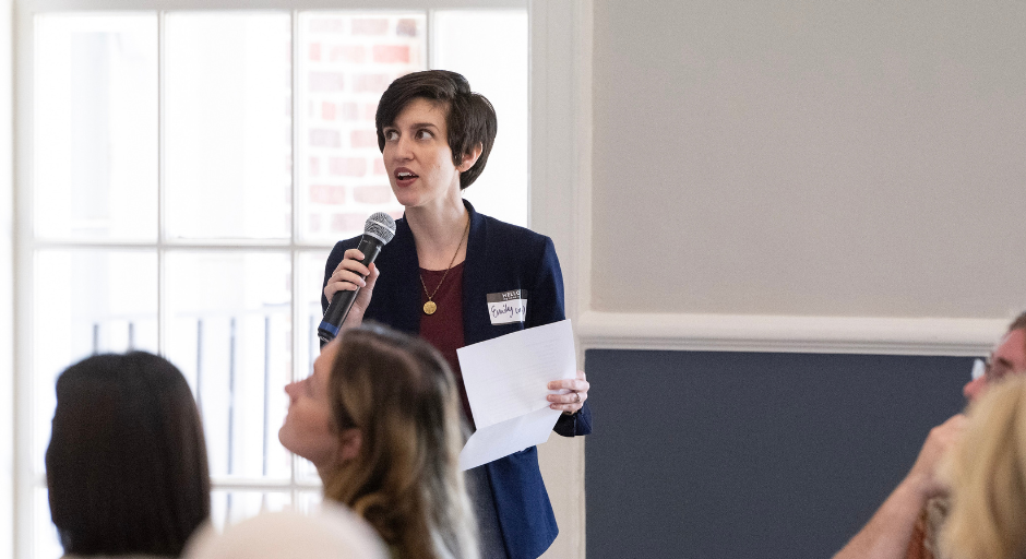 Emily, a white woman with short dark hair, speaks into a microphone during a presentation
