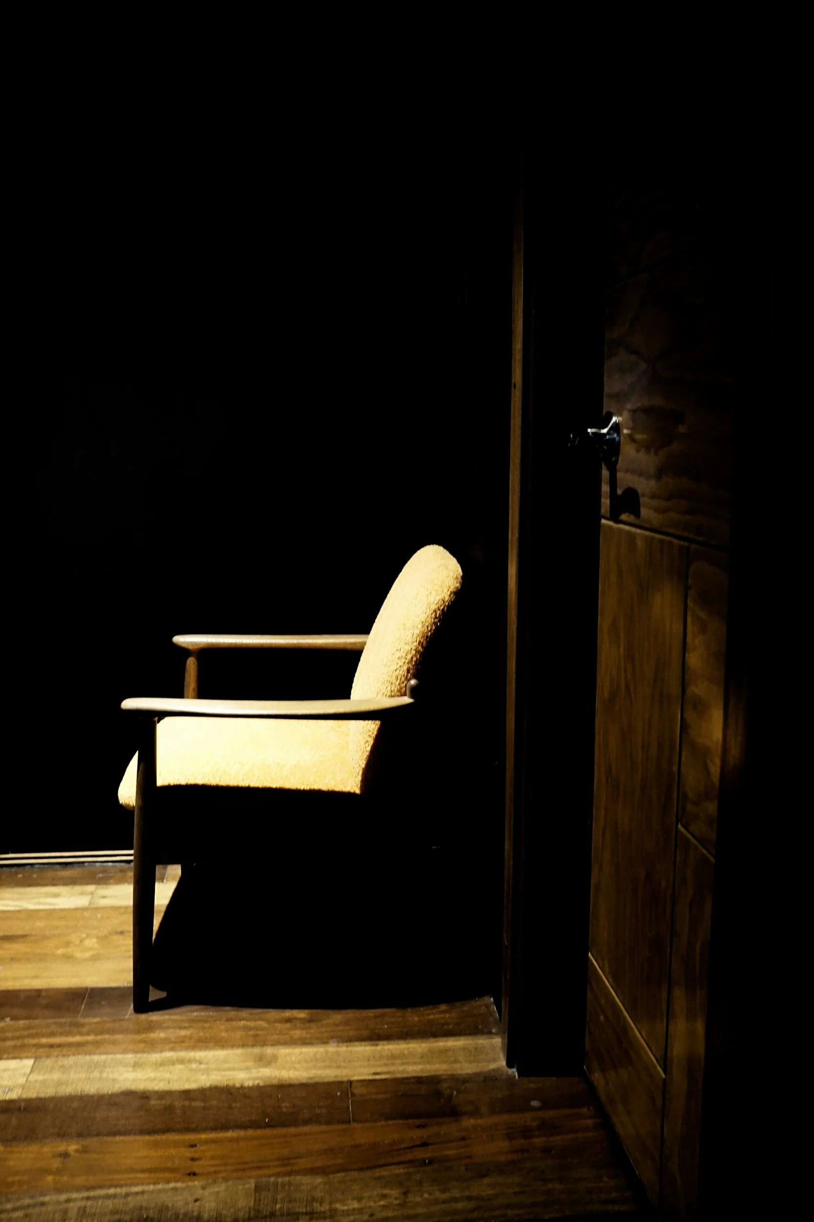 Empty beige armchair with wooden armrests in a dimly lit room with dark walls and wooden flooring.