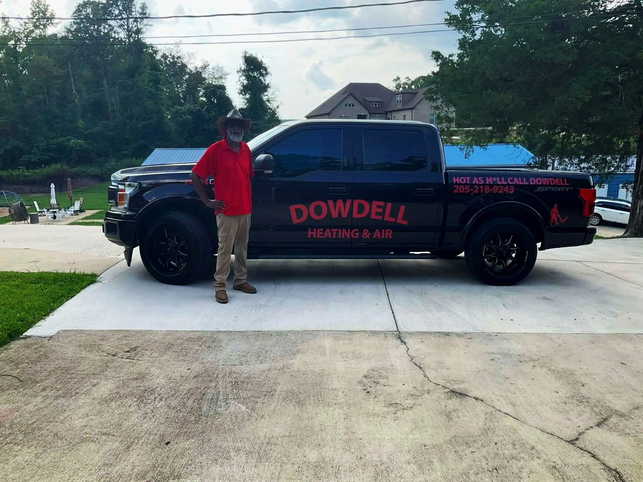 A man stands next to a black pickup truck with red and pink lettering advertising Dowdell Heating & Air, with a phone number. The man is wearing a red shirt, tan pants, and a wide-brimmed hat. In the background, there is a house, trees, and a backyard with outdoor furniture.