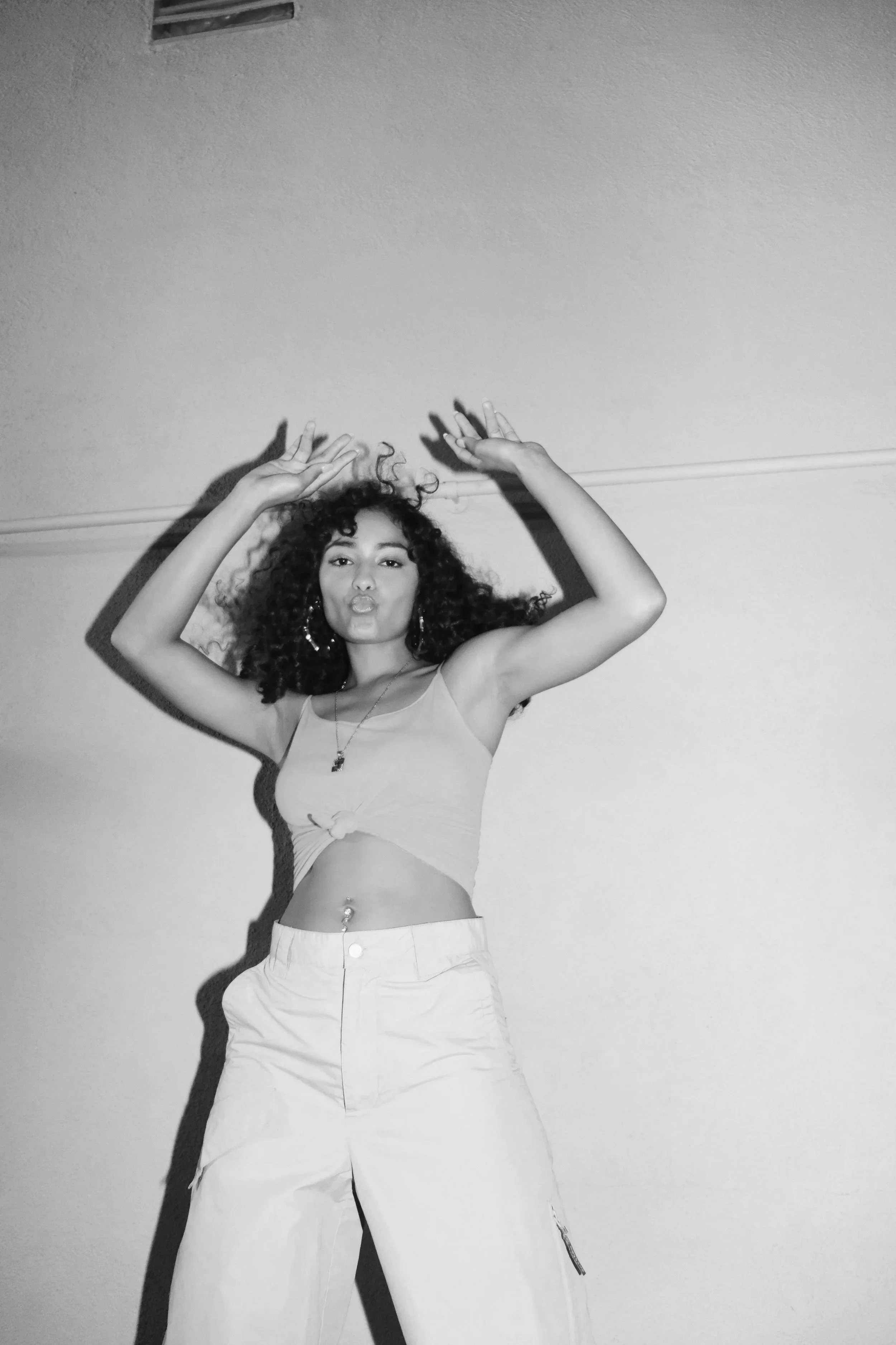 Black and white photo of a woman with curly hair wearing a sleeveless top and high-waisted pants, standing against a plain wall with her arms raised above her head.