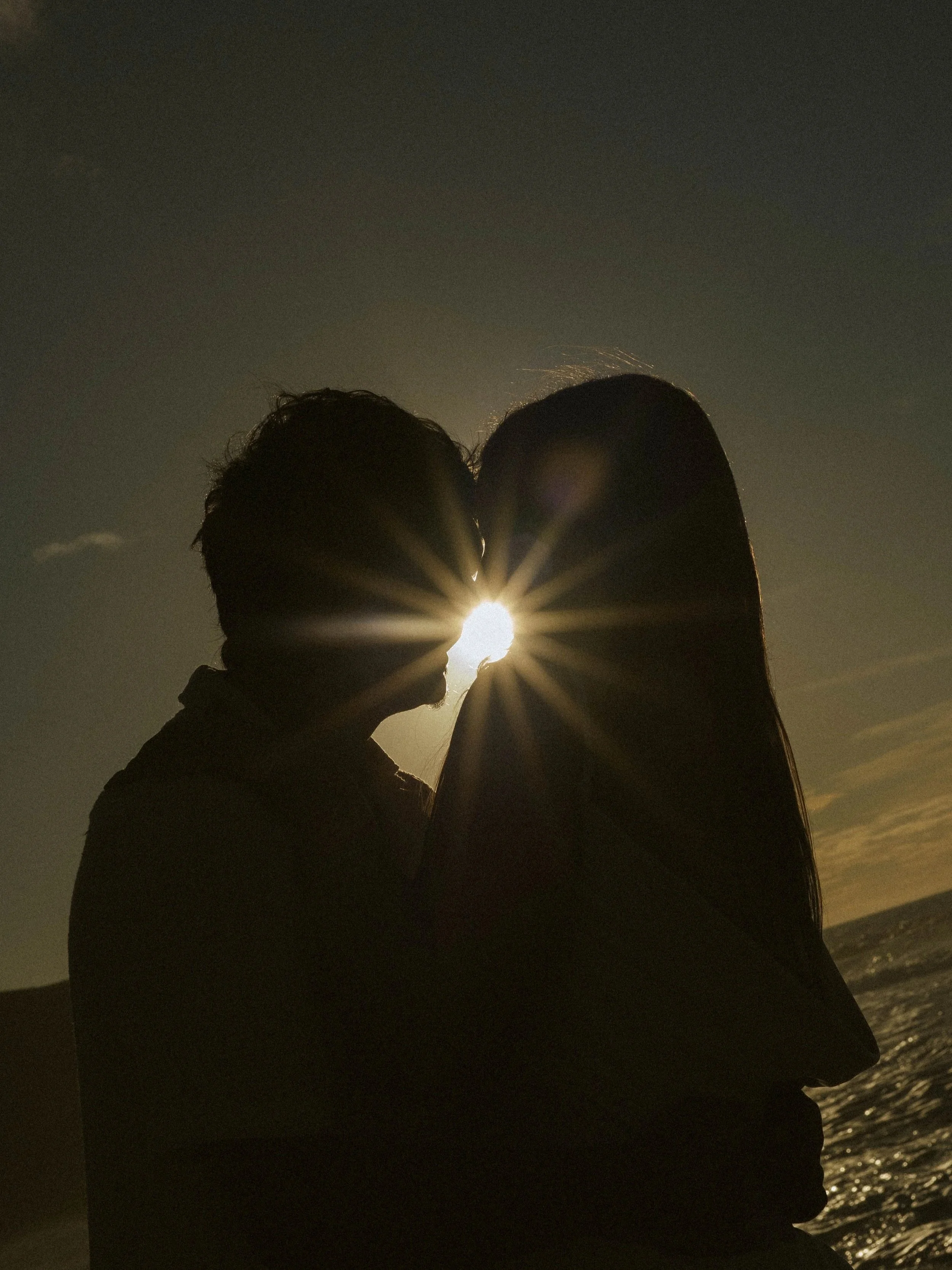 Silhouettes of a couple kissing at sunset near the ocean, with the sun behind them creating a radiant glow and starburst effect.