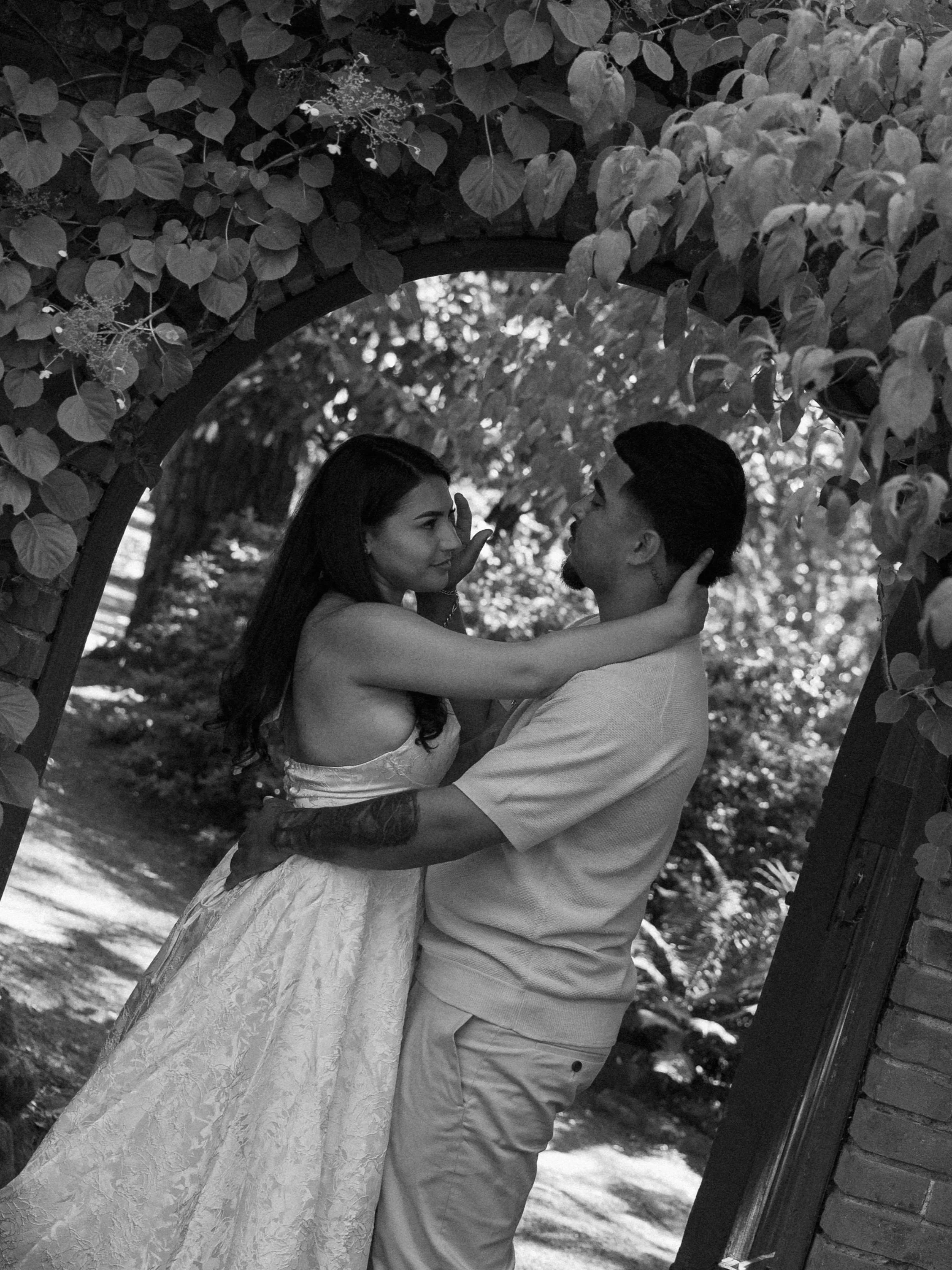 A black and white photo of a couple standing close under a leafy archway, gazing into each other's eyes with affectionate expressions, surrounded by trees and a brick building.