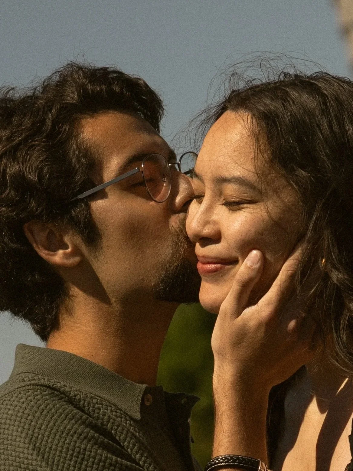 A man with dark hair, glasses, and a goatee is kissing a woman on the cheek. The woman has dark hair, closed eyes, and is smiling softly. The man is gently holding her face, and his other hand is touching her chin.