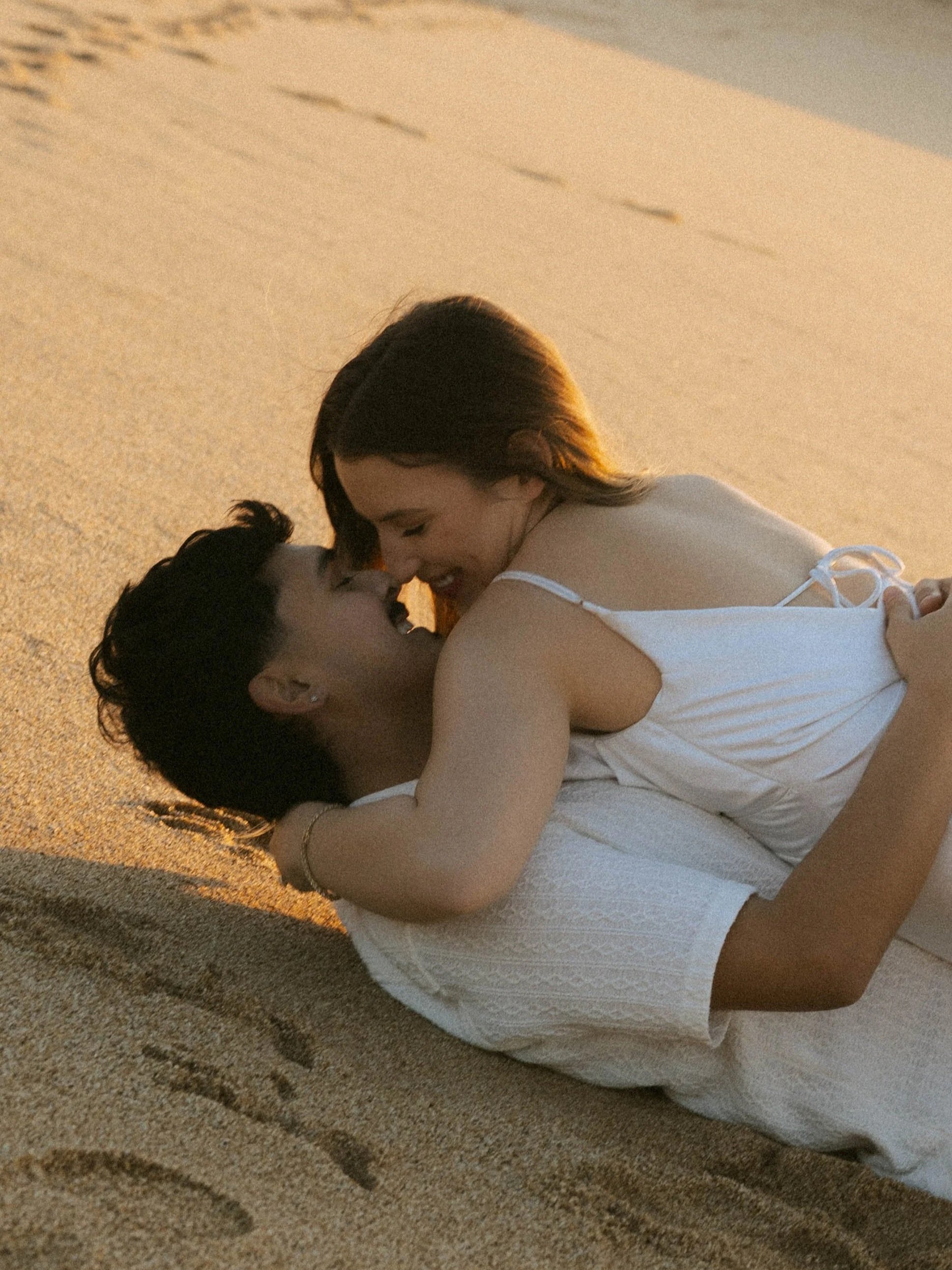 A couple lying on the sandy beach, smiling and close to each other, with the woman's face near the man's face, in warm sunlight.