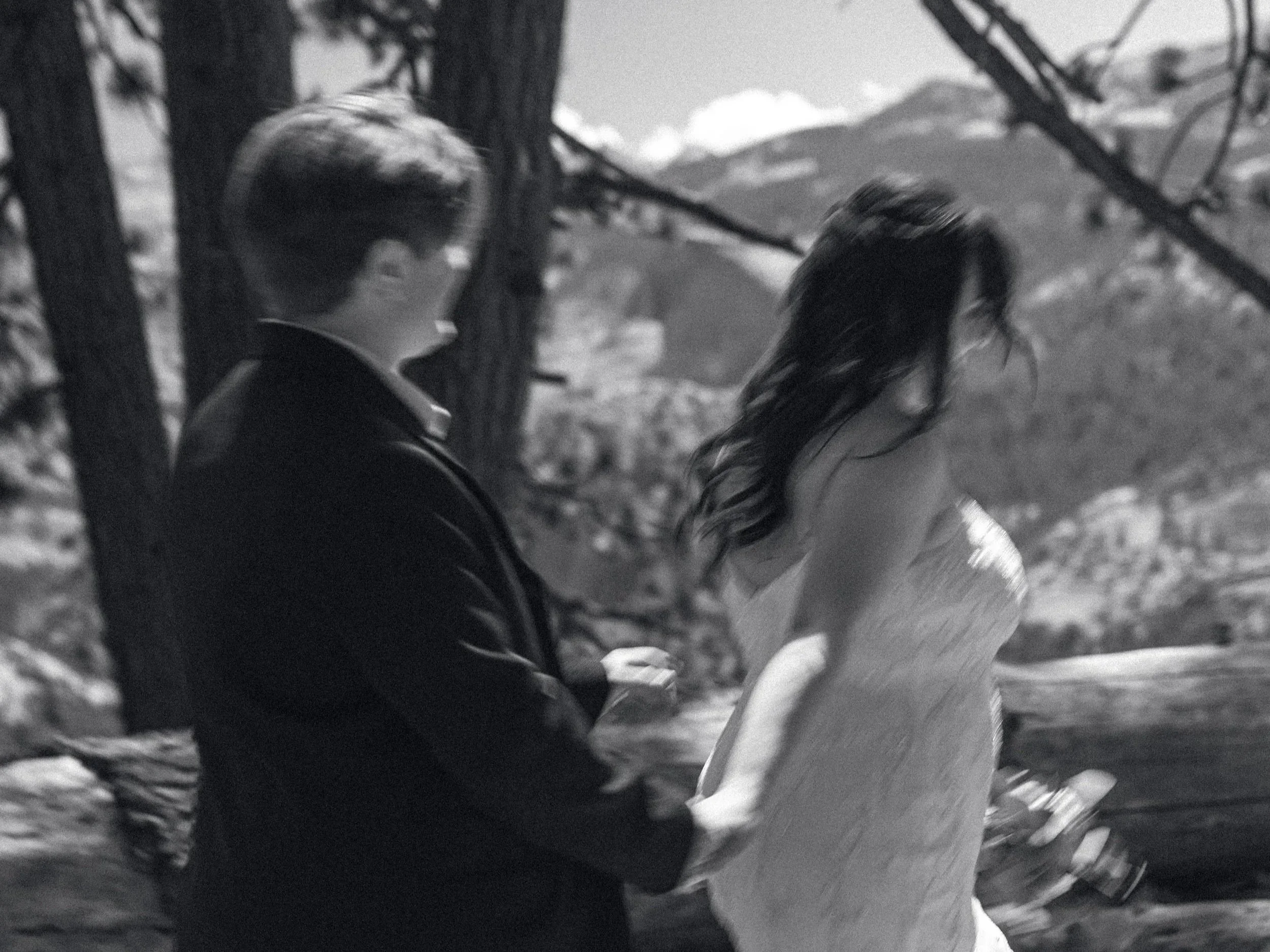 Black and white photo of a couple, with the man sitting on the ground and the woman standing next to him, outdoors in a natural setting with trees and mountains in the background.
