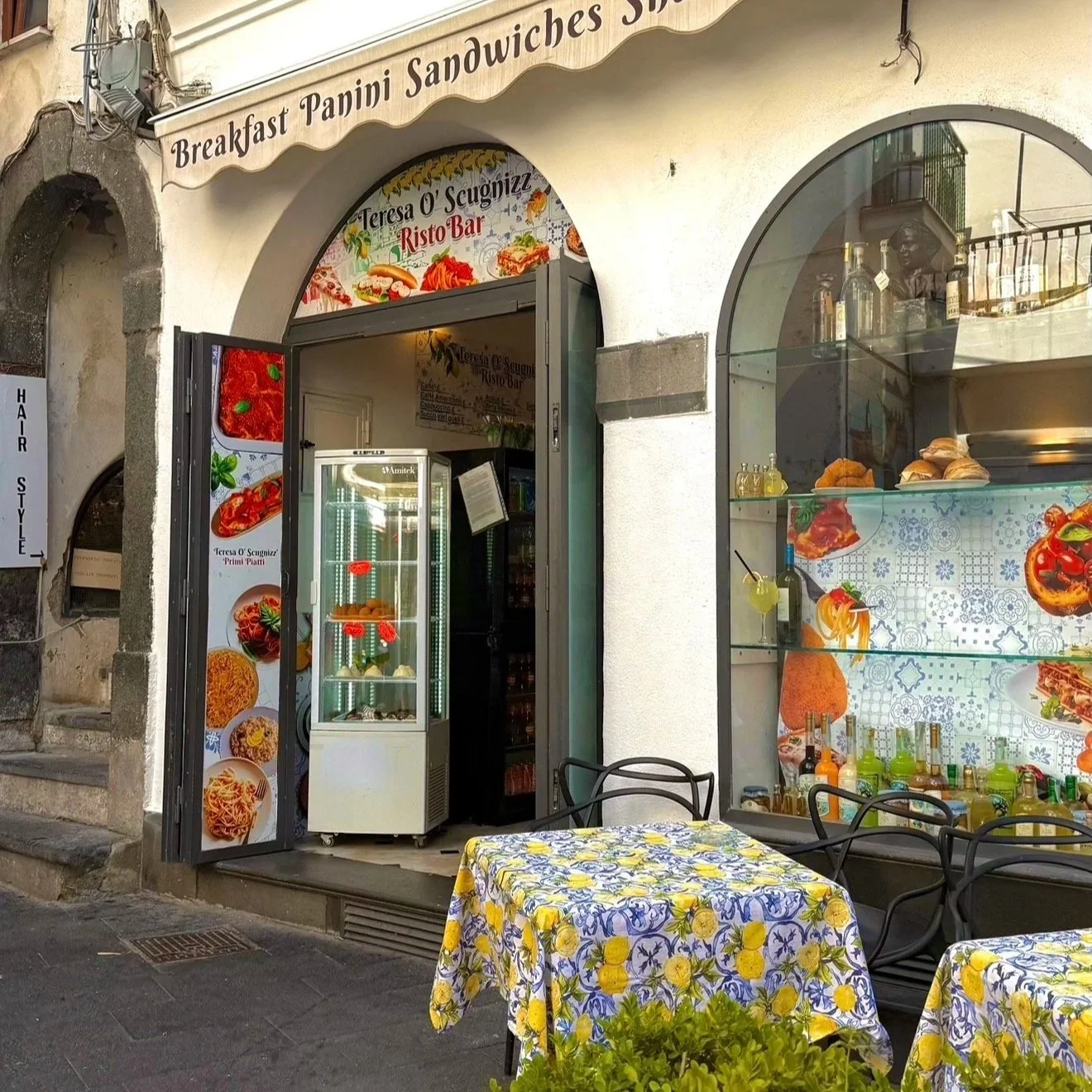 An outdoor view of a small Italian eatery named Teresa O'Scugnizzi Risto Bar with a sign advertising breakfast, panini, and sandwiches. The entrance is open with a display case and colorful pictures of food on the door. There are tables with yellow floral tablecloths and black chairs outside, and a window displaying bottles, pastries, and images of pizza and pasta.