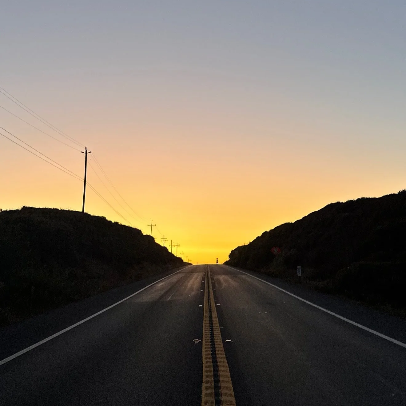 A two-lane road with a yellow dividing line leading toward the horizon at sunset, flanked by dark bushes on both sides and utility poles on the left, with a sky transitioning from fading sunlight to twilight.