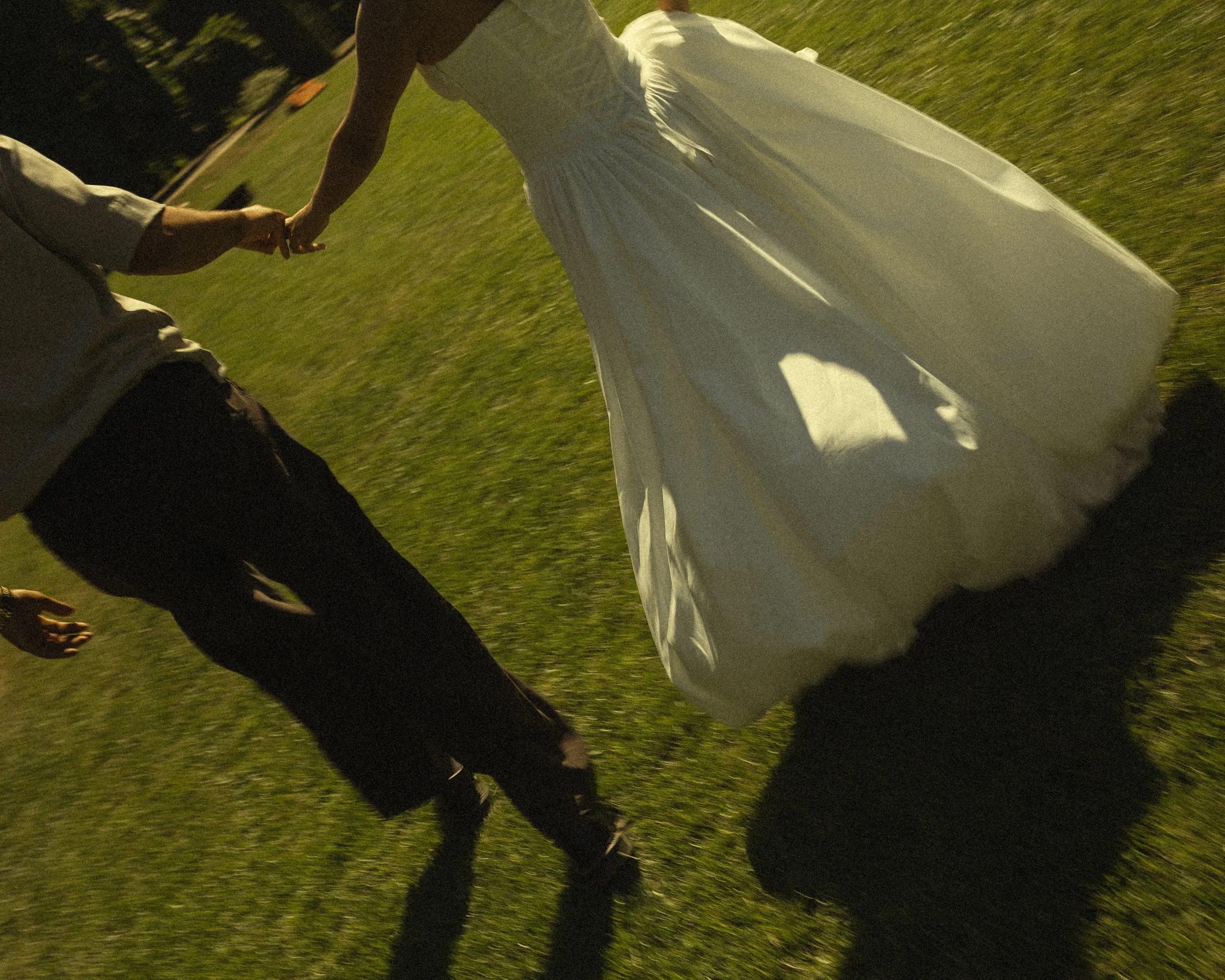 Close-up of a couple holding hands while walking on a grassy field during sunset, with the woman wearing a white dress.