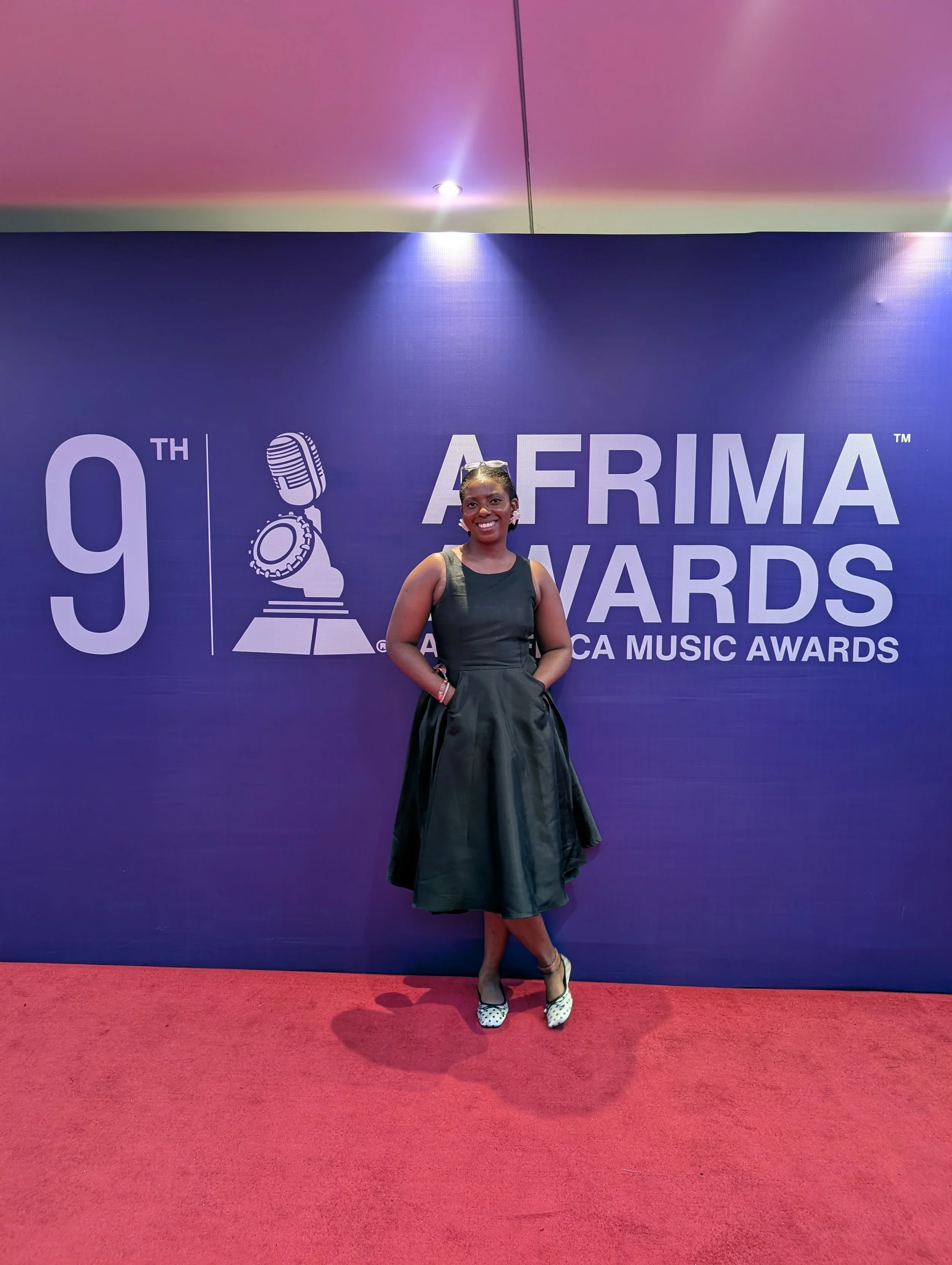A woman in a black dress and polka dot shoes standing on a red carpet in front of a blue backdrop with the text '9th AFIMA Awards'.