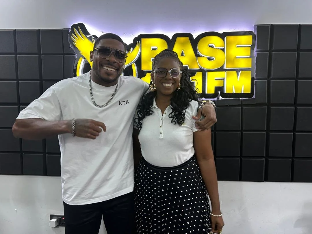 Two people smiling and posing together in front of a black wall with a bright yellow and black sign that says "Base FM". The man is wearing sunglasses, a white t-shirt, a silver chain, and a bracelet, and the woman has curly hair, glasses, a white to