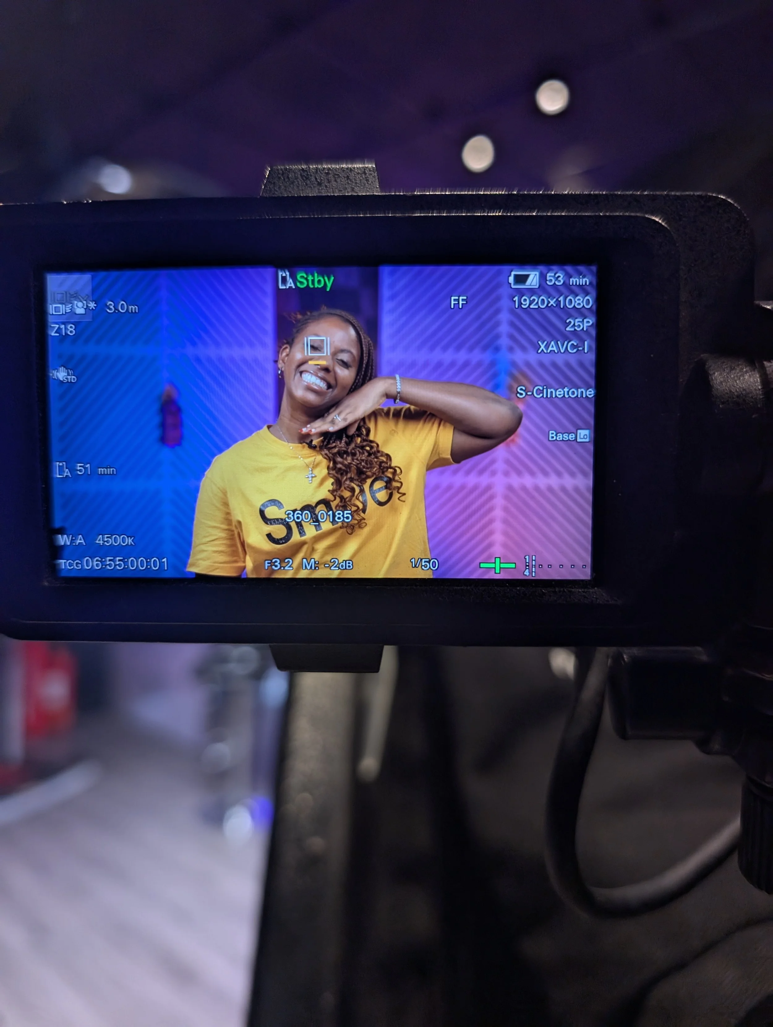 The photo captures the view through a camera's LCD screen or an external monitor showing a smiling woman with long braided hair in a yellow T-shirt, standing in front of a colorful background with purple and red decoration elements, possibly during a
