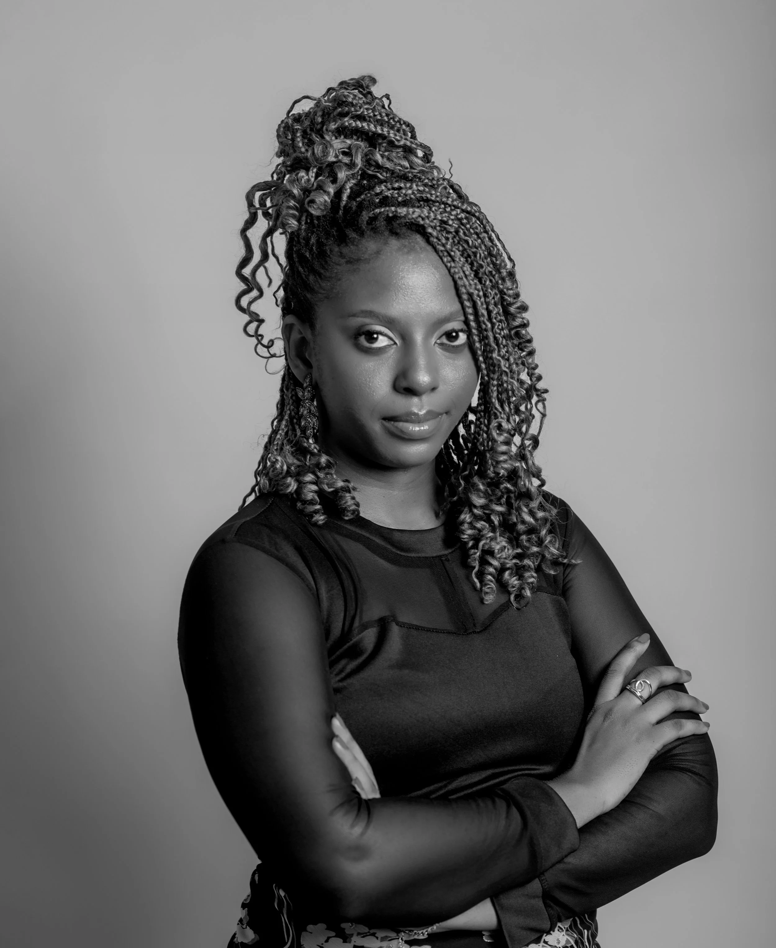 Black-and-white portrait of a woman with curly, styled hair wearing a long-sleeve top and jewelry, looking confidently at the camera with crossed arms.