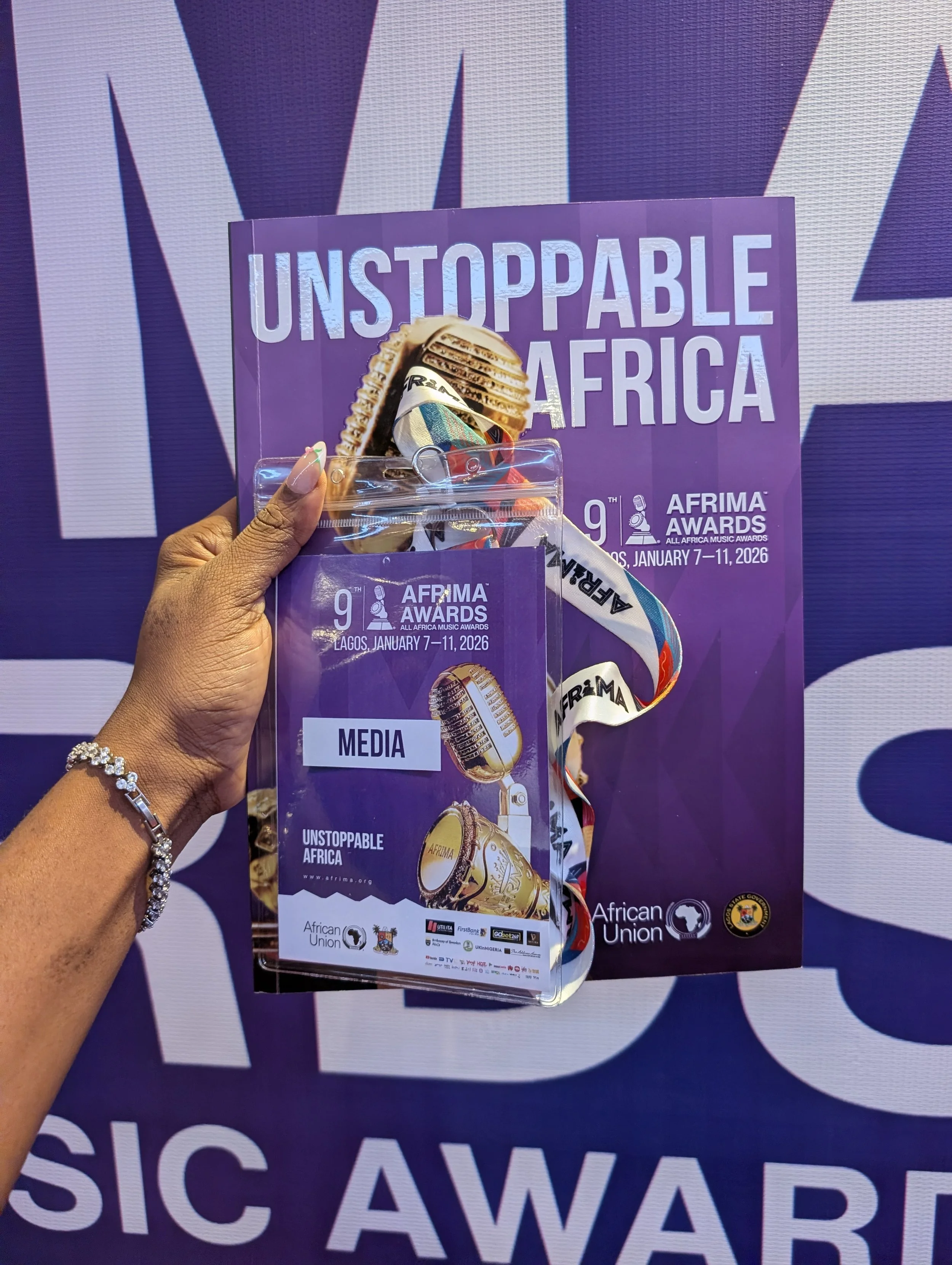 A hand holding a media pass for the AFRIMA Awards 2026 in Lagos, featuring a microphone icon and the word 'MEDIA', with a backdrop displaying 'UNSTOPPABLE AFRICA' and details of the event.
