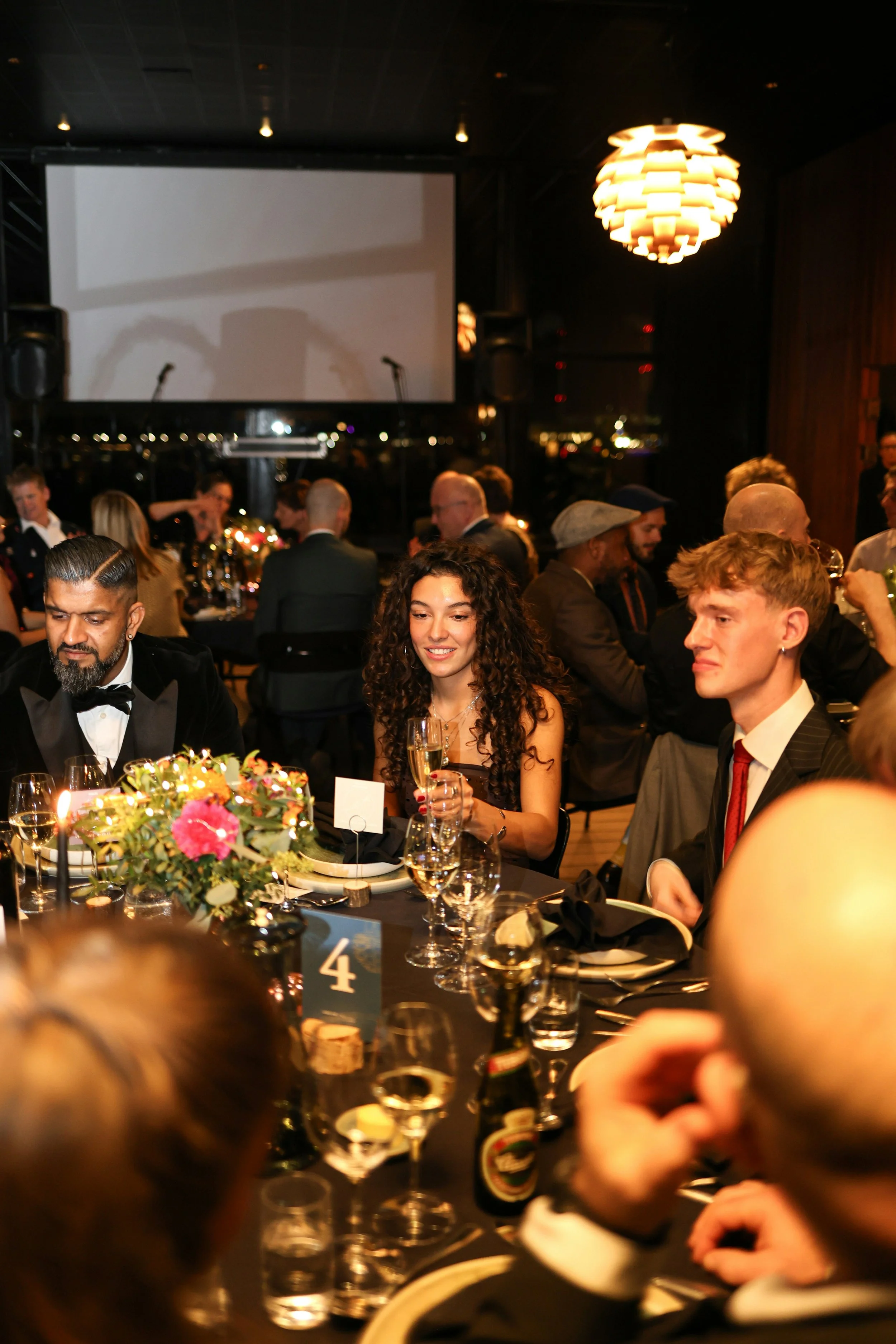 attendees at an event, seated around a table, sharing in conversation and wine, dressed in black tie, with a blurred background of other tables and more attendees