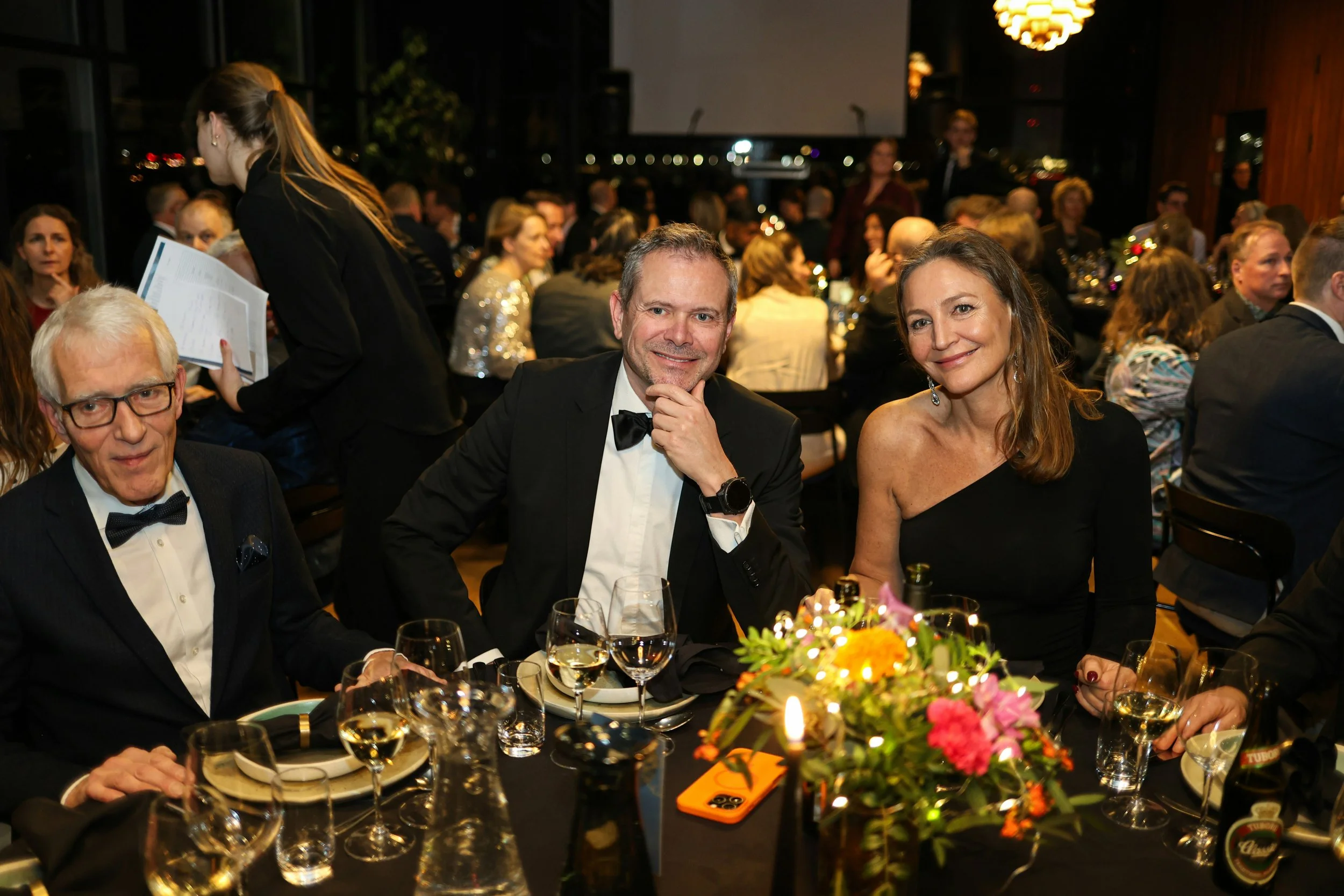 A man in a black suit and woman in black dress sitting at a table at a gala event with a blurred background of gala attendees