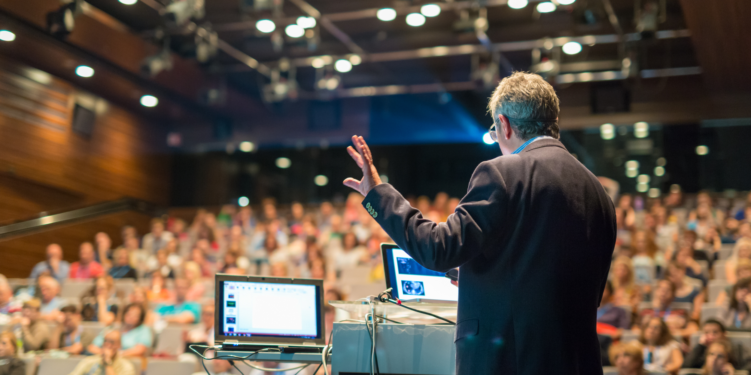 a photo of man, taken from behind, at a podium giving a presentation to a blurred audience