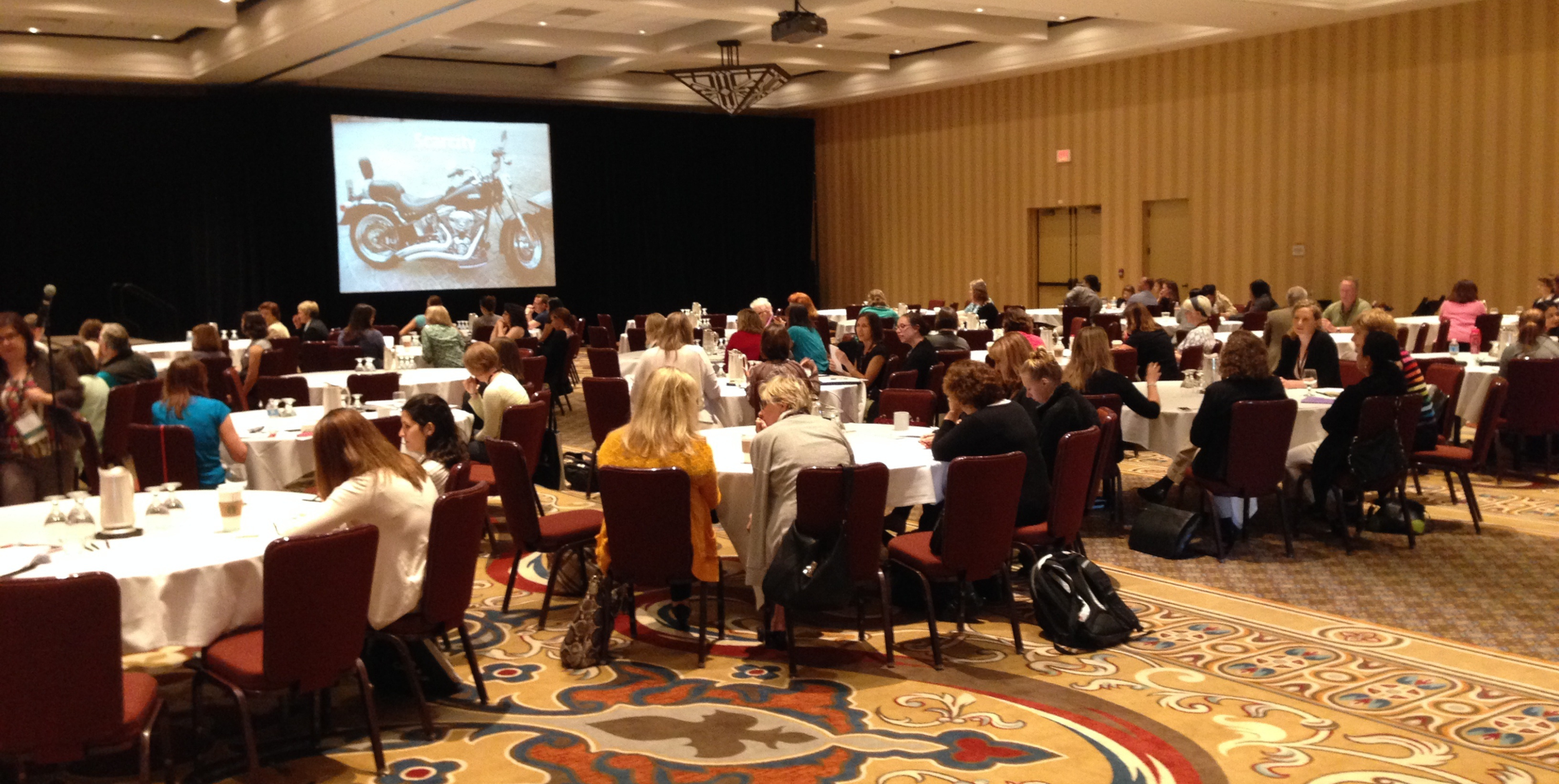 photos of an event with attendees seated at round tables facing a screen which features an image of a  motorcycle
