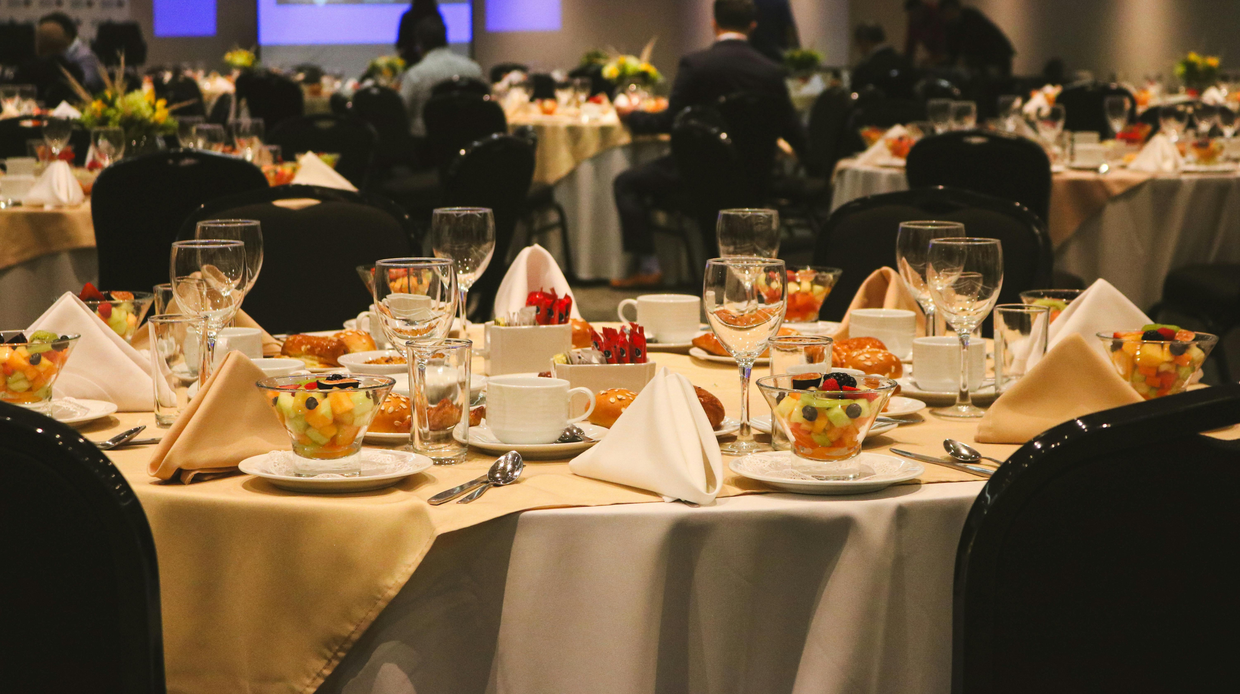 Banquet table set for a meal with glassware, bowls of fruit, pastries, coffee cups, and napkins in a conference or banquet room.