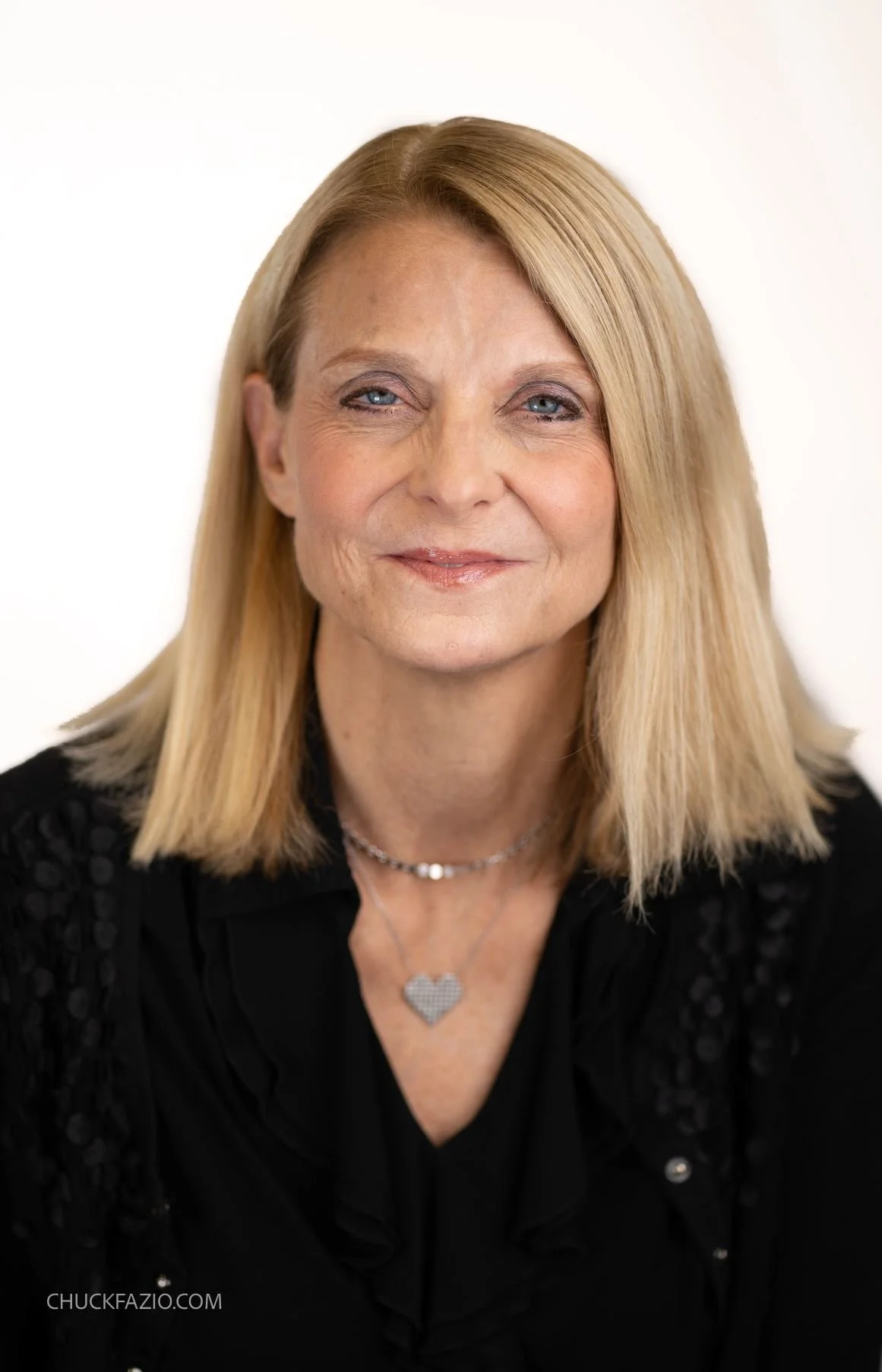 A middle-aged woman with blonde hair and blue eyes, wearing a black top and a silver necklace with a heart-shaped pendant, smiling against a plain white background.
