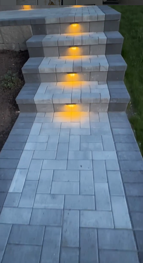 Outdoor stone staircase with built-in yellow LED lights illuminating each step, leading to a higher level, with grass visible on the right side.