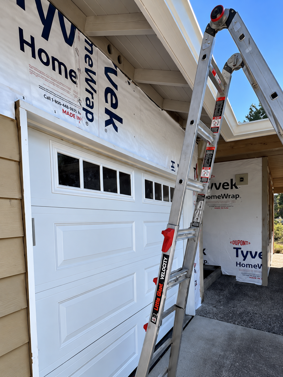 Construction site showing a garage door with window panels, a ladder, and house wrap with brand labels, under a partially built roof with roofing shingles.