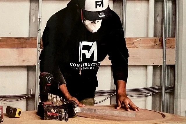 Person working on a woodworking project in a workshop, wearing a black hoodie with a logo, a black cap, and a face mask, using a cordless drill on a wood piece.