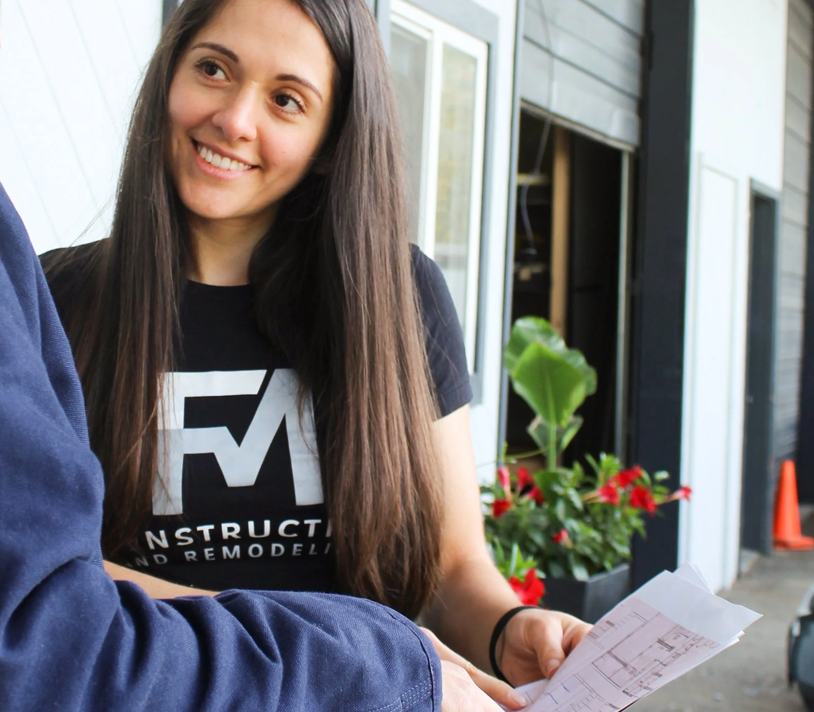 A woman with long brown hair smiling while talking to someone outdoors, holding a sheet of paper with architectural plans. She is wearing a black T-shirt with white text that says, 'CONSTRUCT, BUILD, REMODEL.' In the background, there are green plants, red flowers, and part of a building with windows and siding.