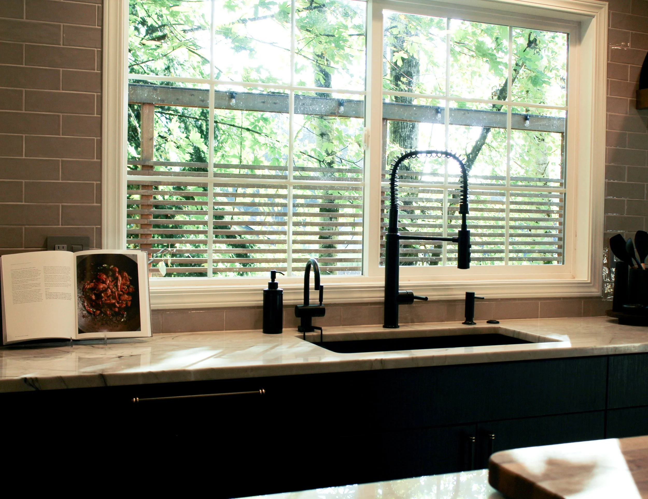 Kitchen window with white frame, view of green trees outside, black kitchen faucet, open cookbook on marble countertop, gray brick backsplash.