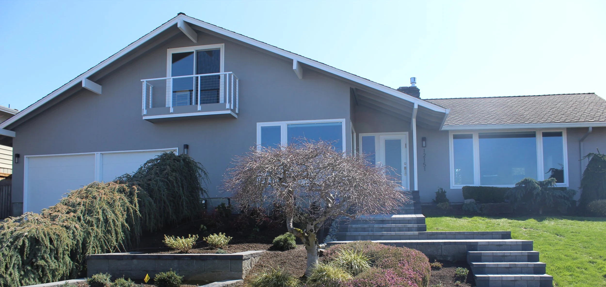 A modern two-story house with a gray exterior, large front windows, and a small balcony on the upper level. The house features a sloped roof and a front entrance with stone steps and landscaping, including a small tree and shrubbery in the yard.