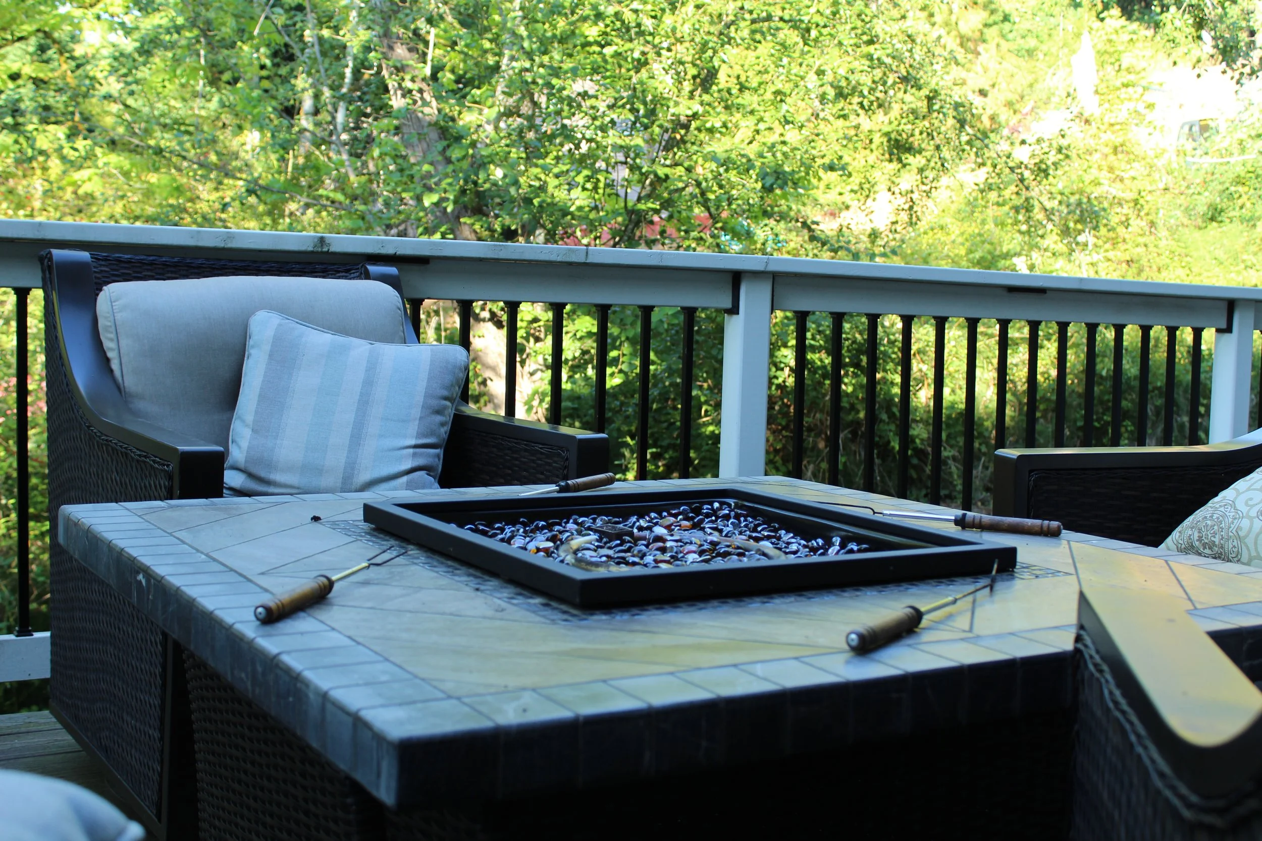 Outdoor patio table with a tray of marbles, surrounded by wicker chairs with cushions, overlooking trees and greenery in the background.