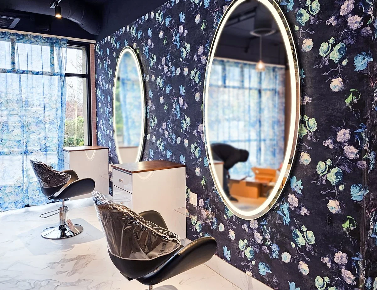 A hair salon with two black styling chairs in front of a dark blue floral wallpapered wall, featuring one large oval mirror with a backlit frame, and a window with sheer blue floral curtains.