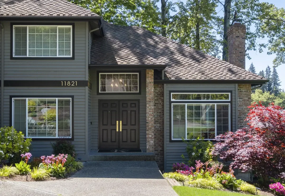 Front view of a gray two-story house with large windows, a black front door with gold handles, a brick chimney, and landscaped garden with colorful flowers and shrubs.