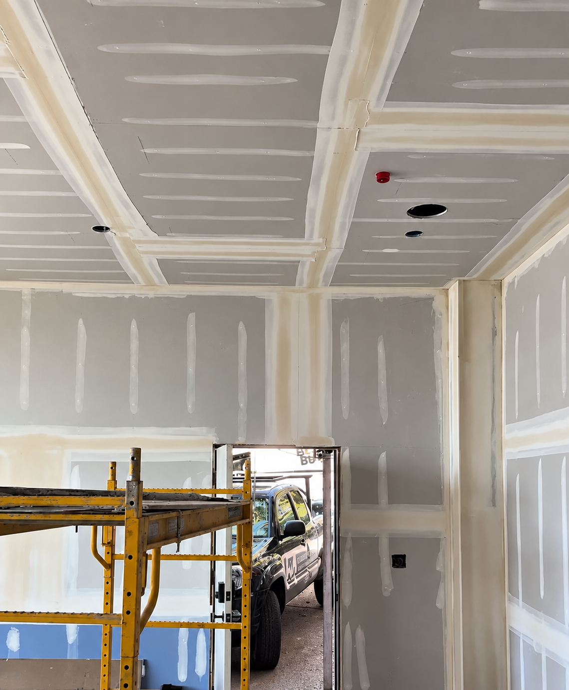 Interior of a room under construction with drywall installed, taped, and mudded, a yellow scaffold in the foreground, and a vehicle visible through an open doorway.