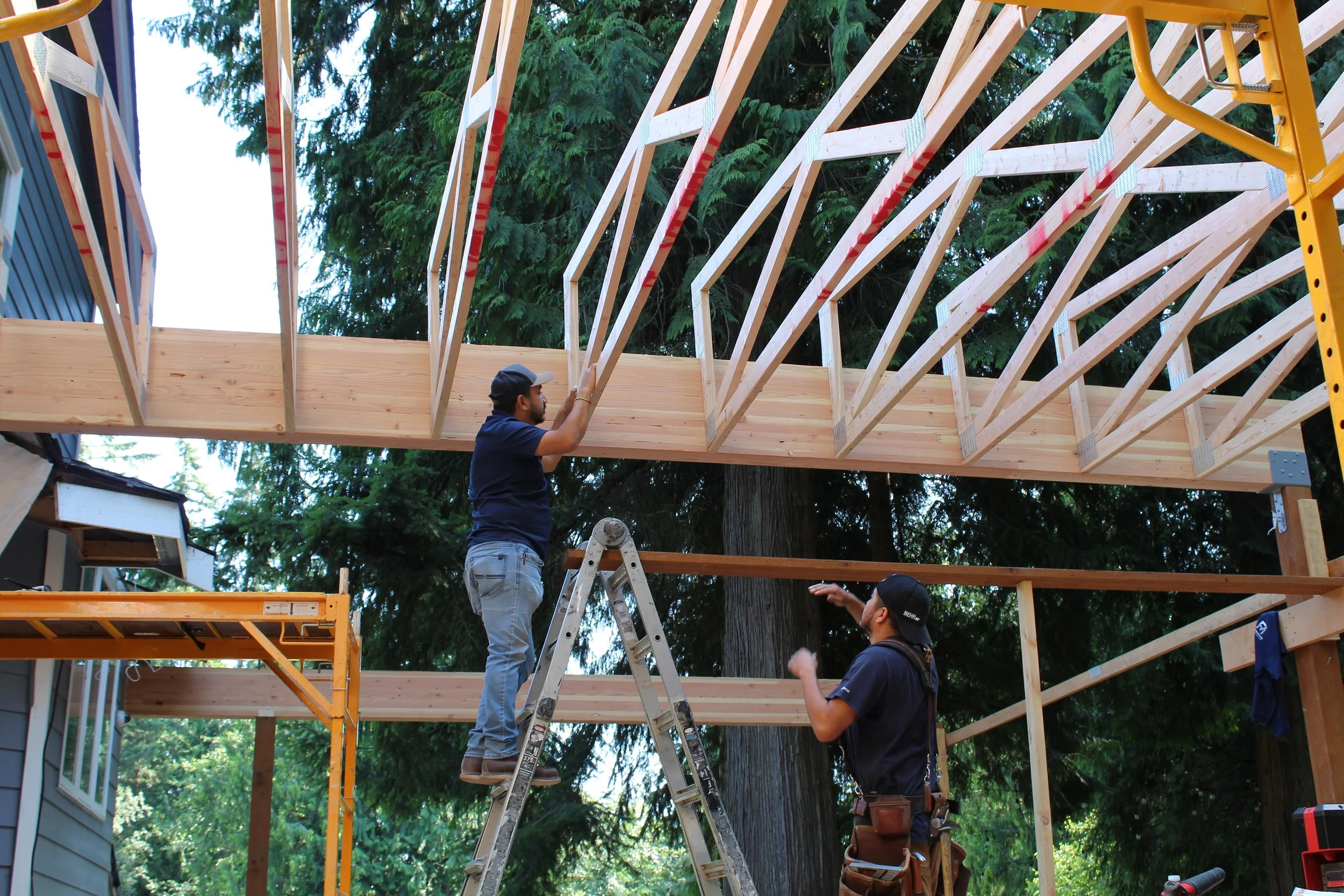 Workers constructing a wooden frame for a house, one standing on a ladder and another on the ground, with trees in the background.
