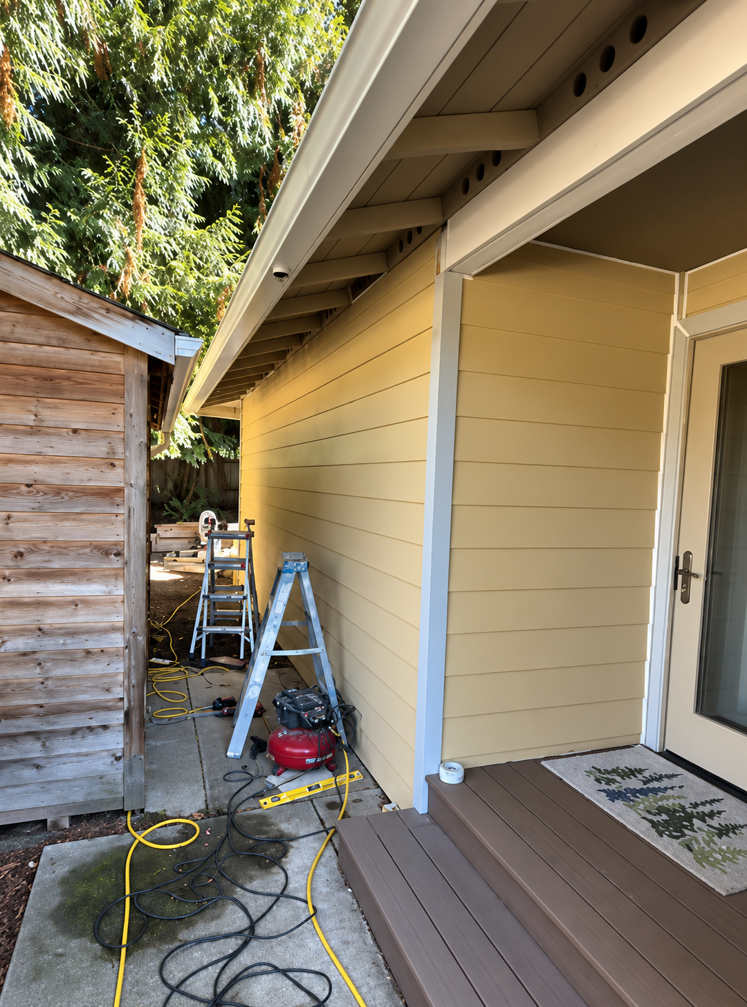 Side view of a house with yellow siding, a wooden deck with seating, and a garden shed in the background, with construction tools and cords on the ground.