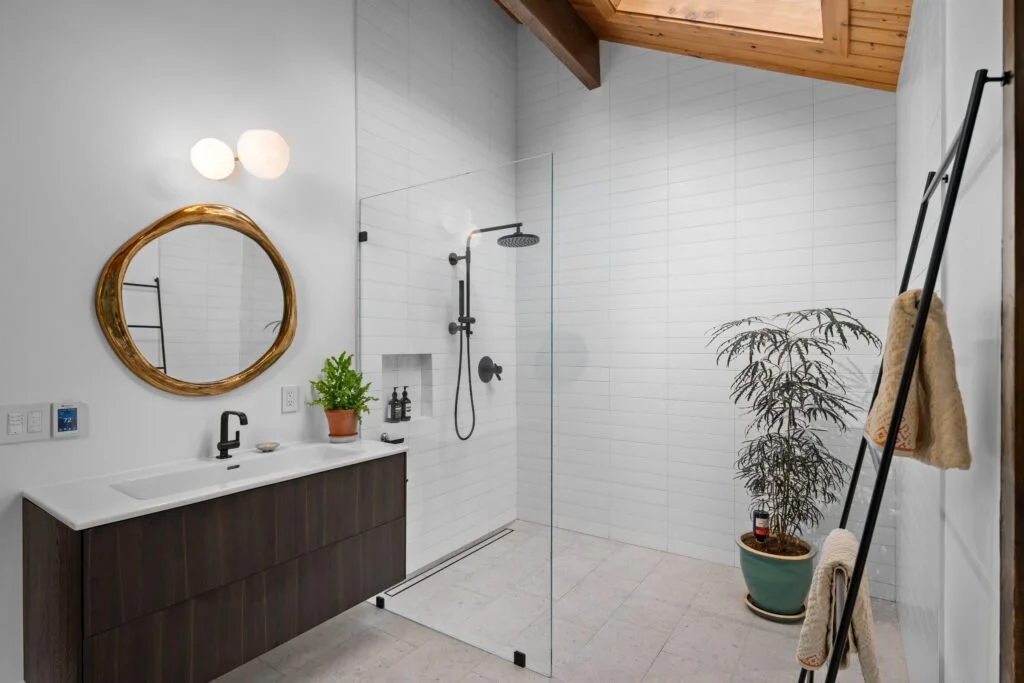 Modern bathroom featuring a vanity with a mirror, a potted plant, and a shower area with a rainfall showerhead separated by a glass panel. There are also two potted plants, a ladder with towels, and a wooden ceiling.