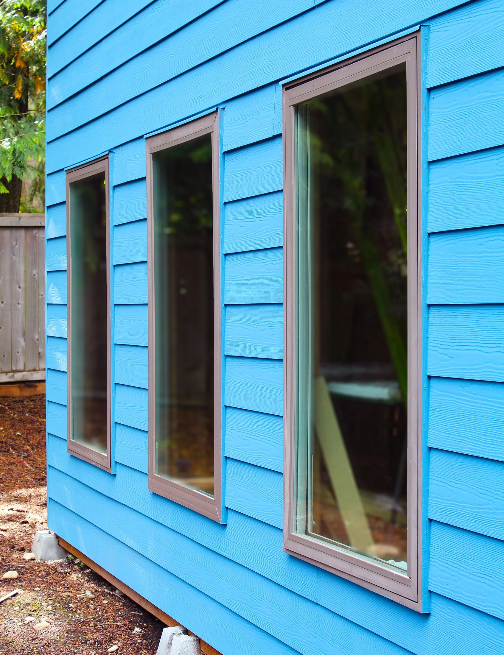 Close-up of a bright blue house exterior with three large rectangular windows with brown frames. The house is elevated on concrete blocks, and part of a wooden fence and green trees are visible in the background.
