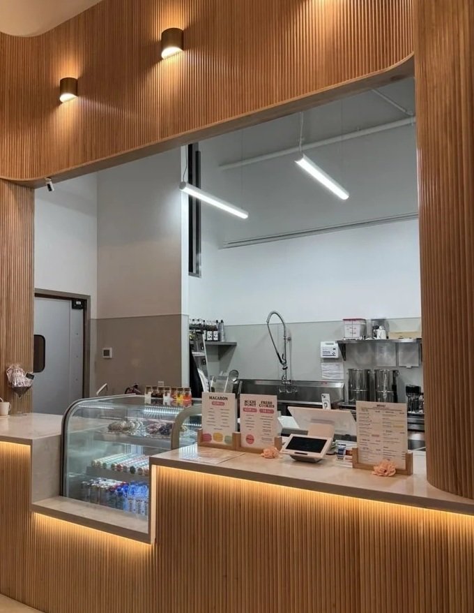 Interior of a cafe or bakery with a wooden counter, menu displays, and a transparent display case with bottled drinks.