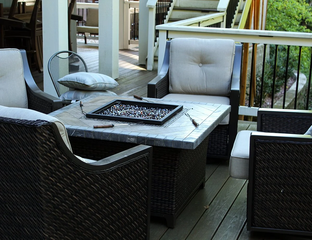 Outdoor patio with a square coffee table, wicker chairs with cushions, a glass beaded game on the table, and a pillow stacked on a chair, surrounded by wooden railing and stairs.