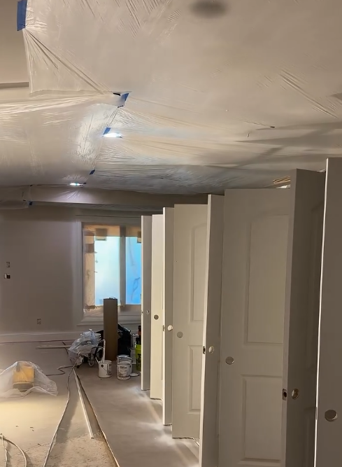 Interior of a house under construction or renovation, with partially installed doors, plastic sheeting on the ceiling, and construction supplies on the floor.