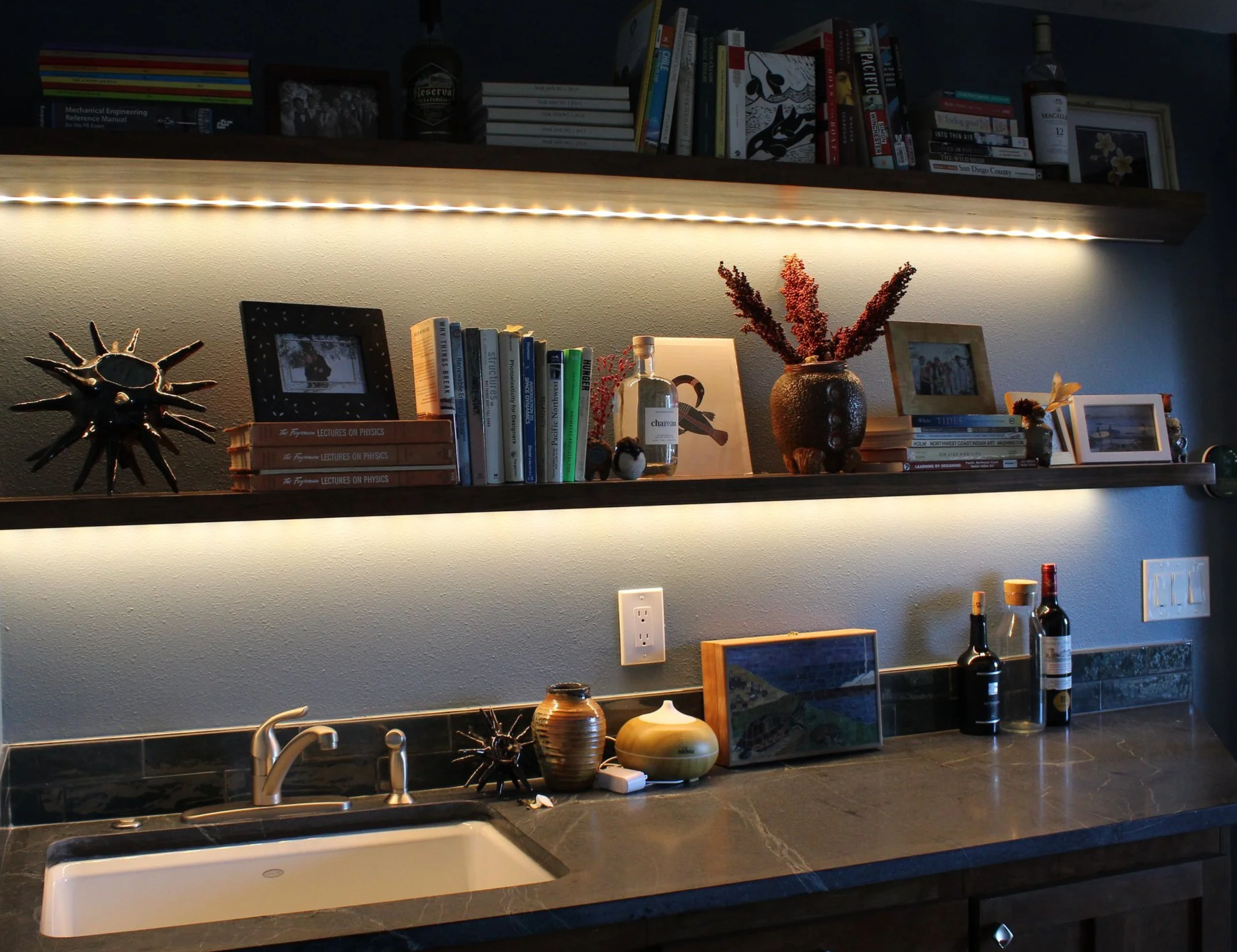 A kitchen with a dark countertop, a white sink, and a backsplash with shelves above there are multiple decorative items, books, framed photos, vases with dried flowers, and bottles of alcohol. The shelves have lighting underneath. Various kitchen utensils and jars are on the countertop.