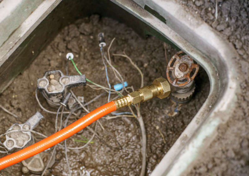 Close-up of a plumbing repair in a sink, showing an orange flexible water supply hose connected to a brass valve, with exposed pipes and wiring in a sink basin.