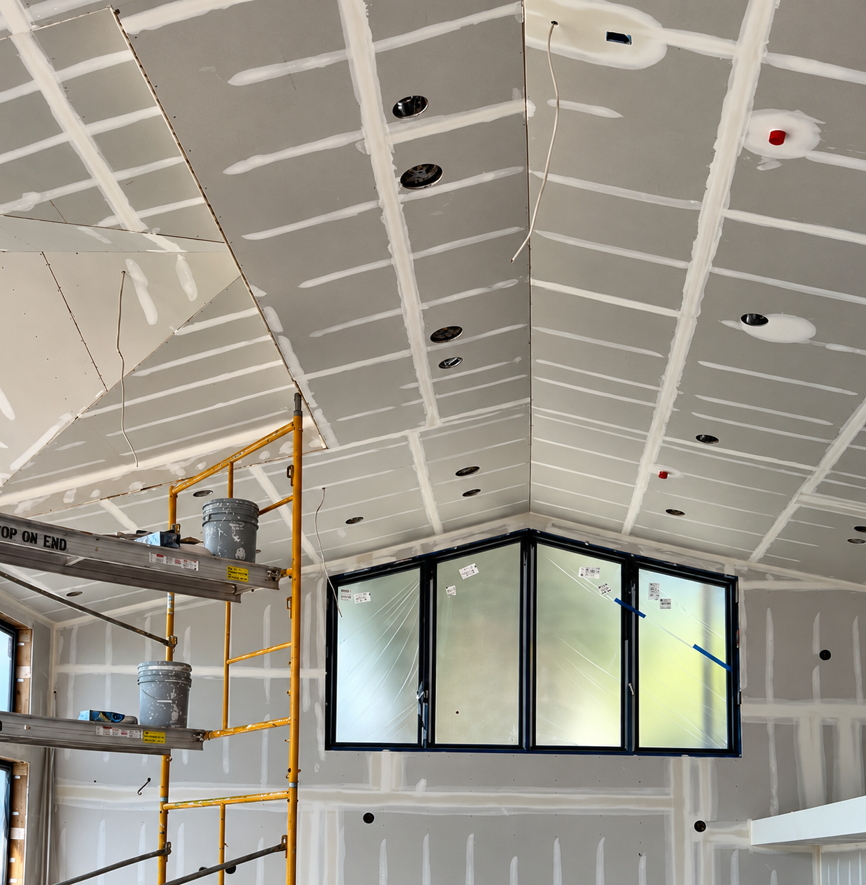Interior construction site with drywall installation on vaulted ceiling, with visible screw holes and joint compound, and a large triangular window in the corner.
